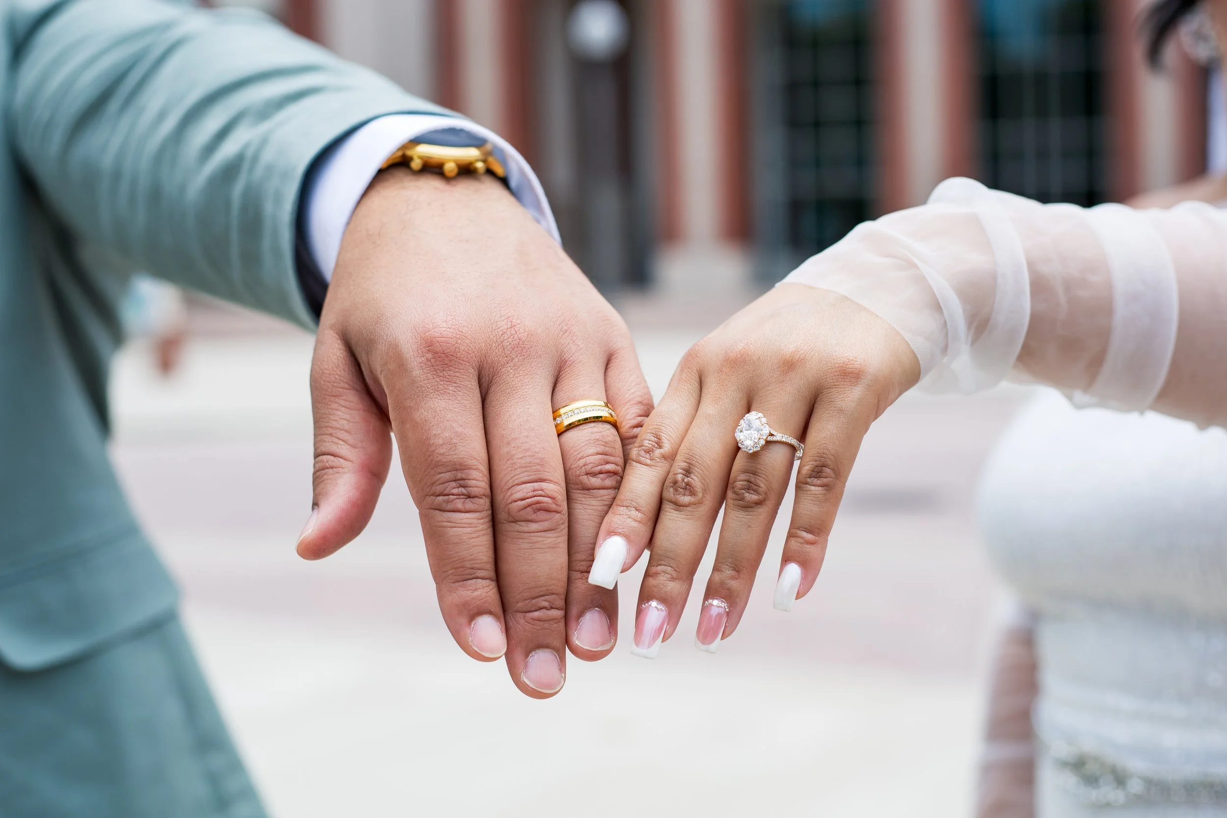Close-up of a couple showing their wedding rings on their ring fingers, with the bride's ring featuring a large diamond and the groom's band being gold, both with their hands touching outdoors.