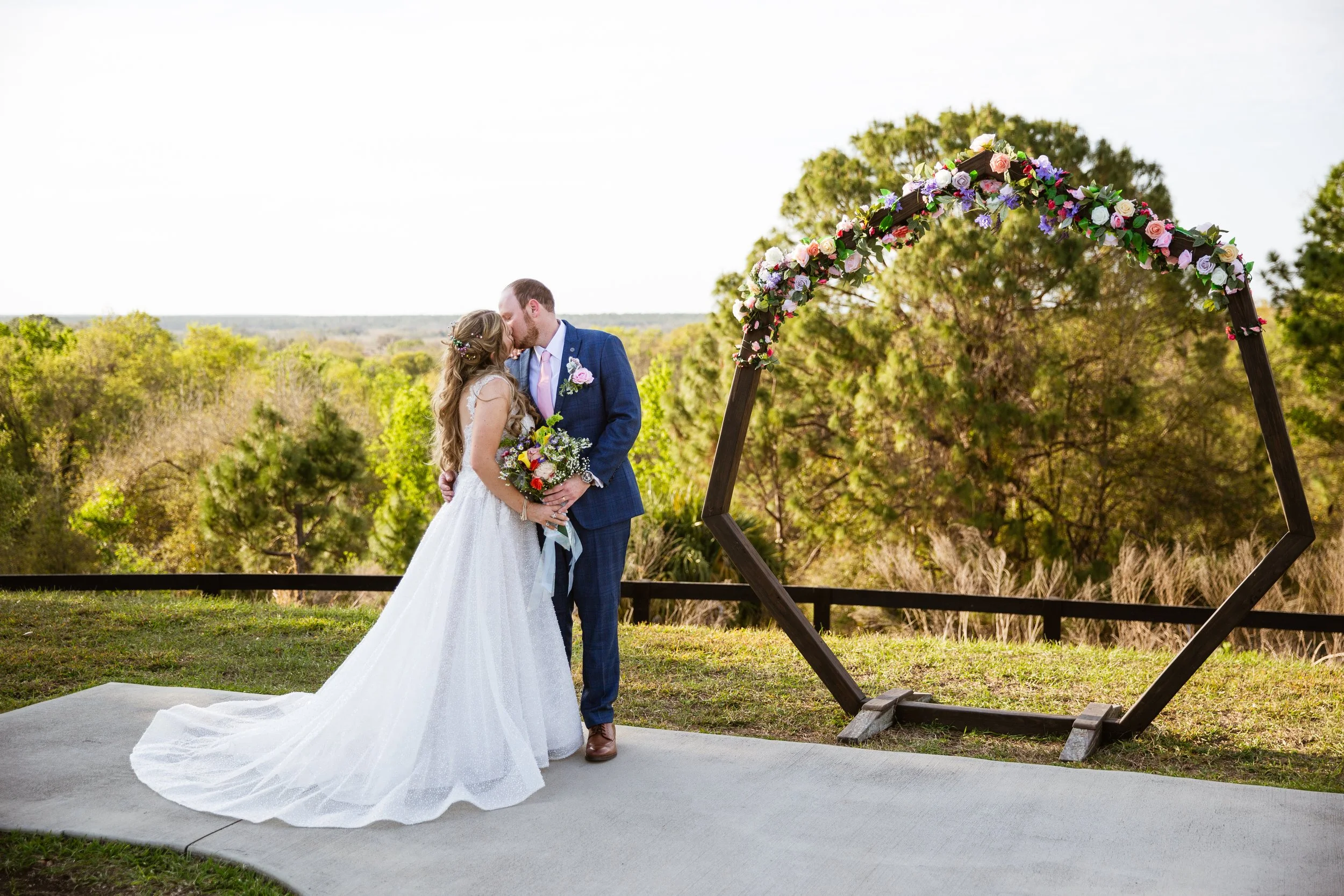 A bride and groom share a kiss during their outdoor wedding ceremony, standing on a small platform in front of a geometric floral arch with trees and greenery in the background.