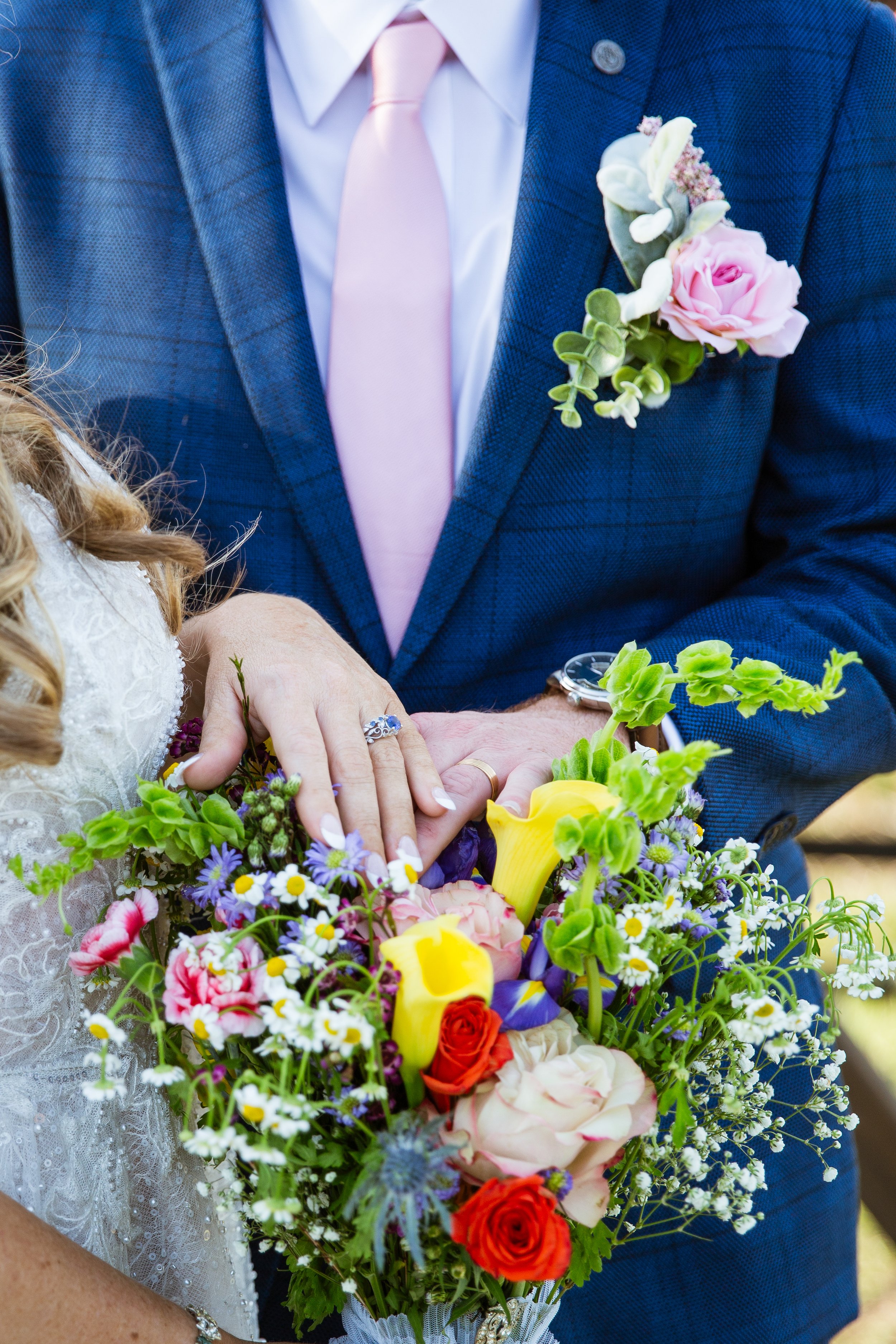 Close-up of a bride and groom holding a vibrant bouquet of flowers during their wedding ceremony, with the groom wearing a blue suit, pink tie, and boutonniere, and the bride showing an engagement ring and wedding band.