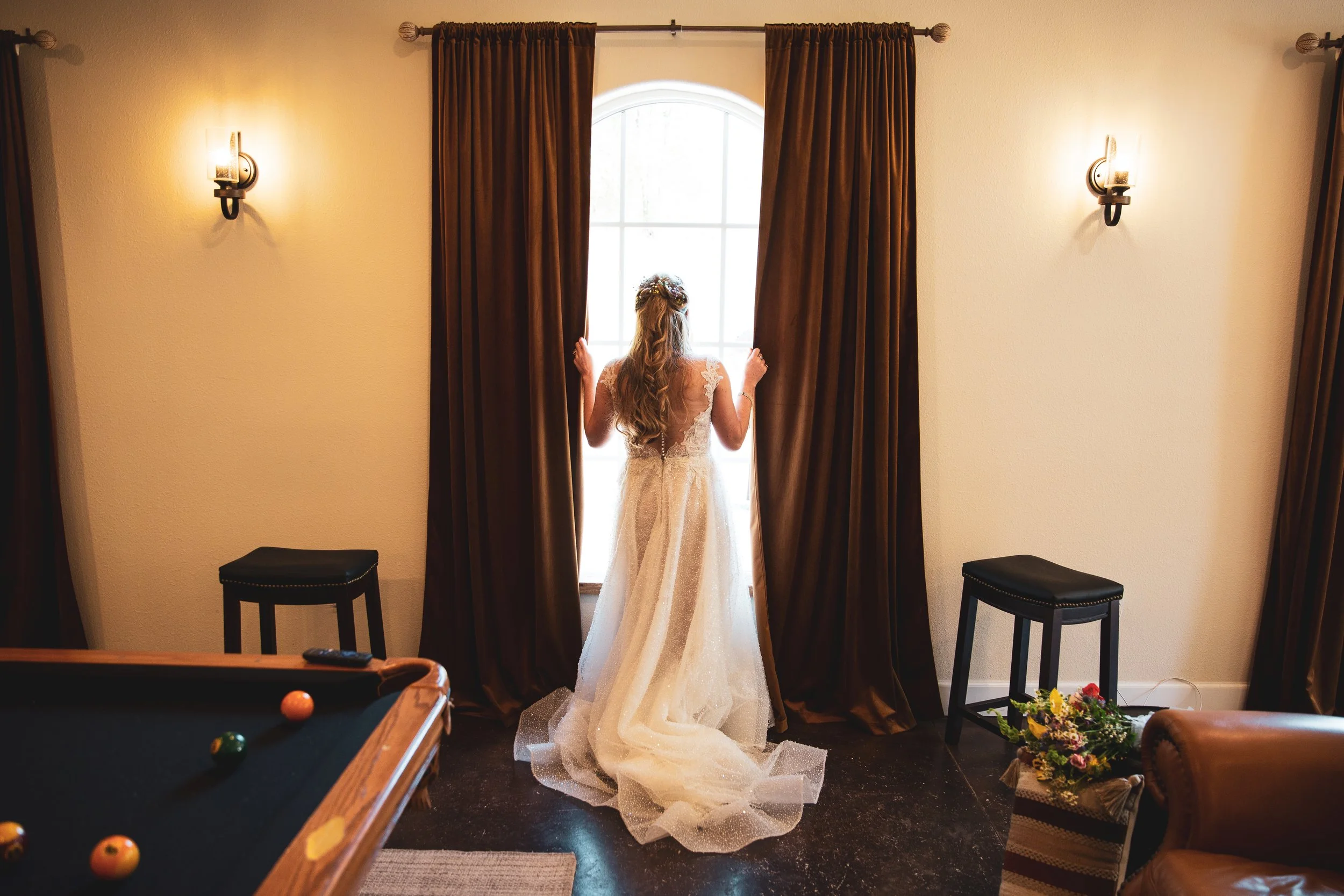 A woman in a wedding dress standing by a large window, opening brown curtains, with her back to the camera.