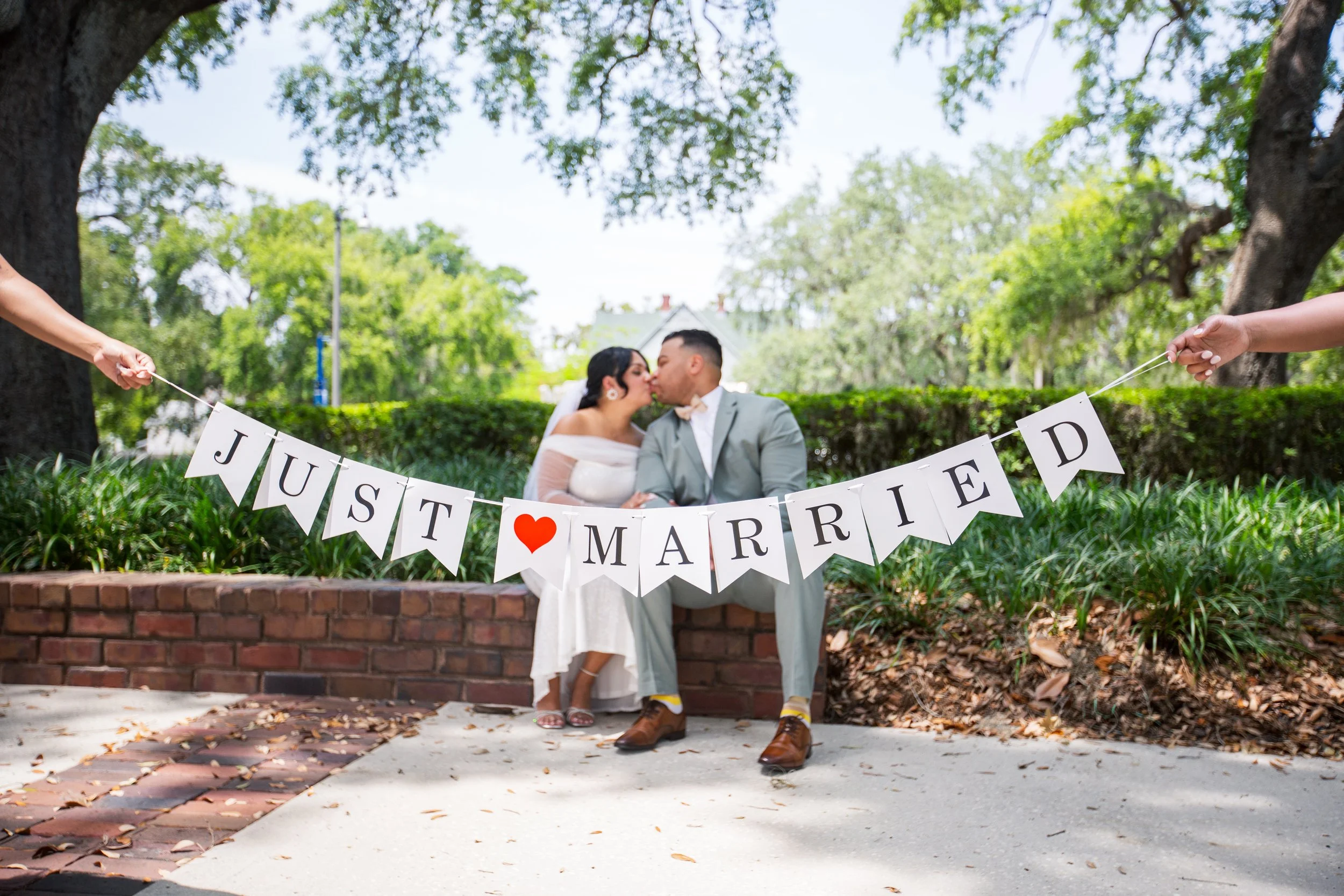 A bride and groom sitting on a low brick wall under a large tree, sharing a kiss. They are holding a white banner with black letters spelling 'JUST MARRIED' with a red heart in the middle, held by two people on either side. The scene is outdoors with greenery and a house visible in the background.