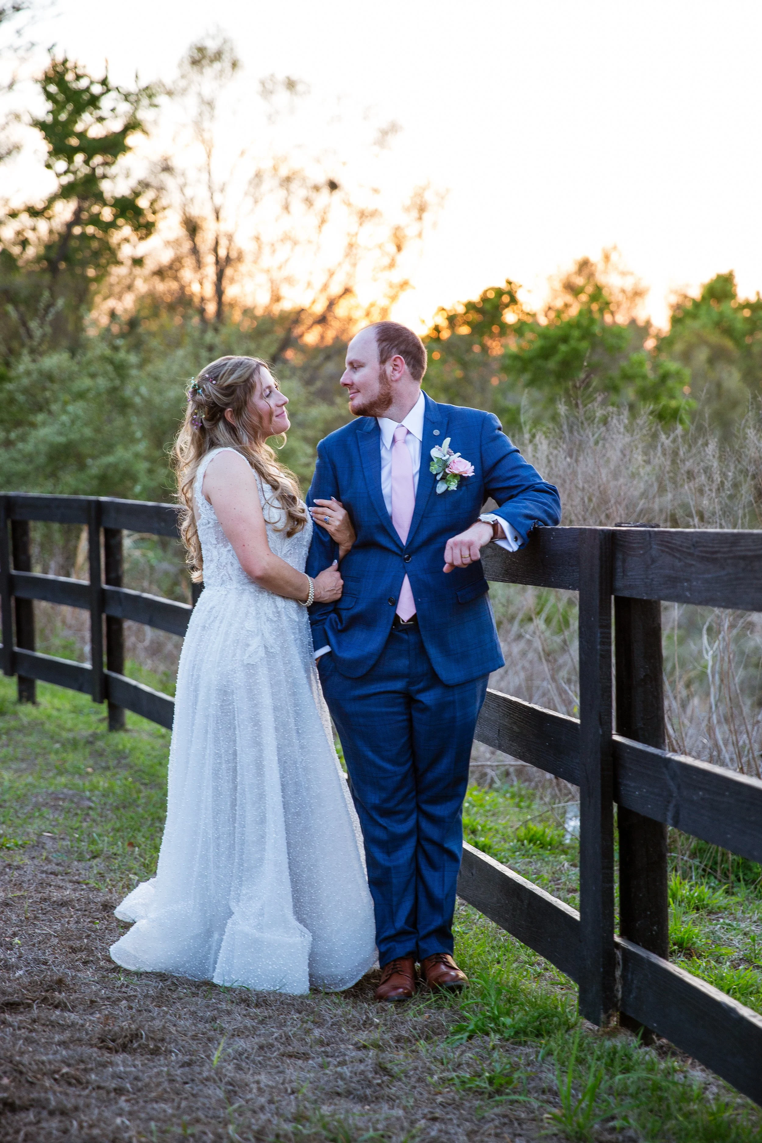 A bride and groom stand outdoors at sunset, gazing at each other near a wooden fence with trees and bushes in the background, during their wedding photo.