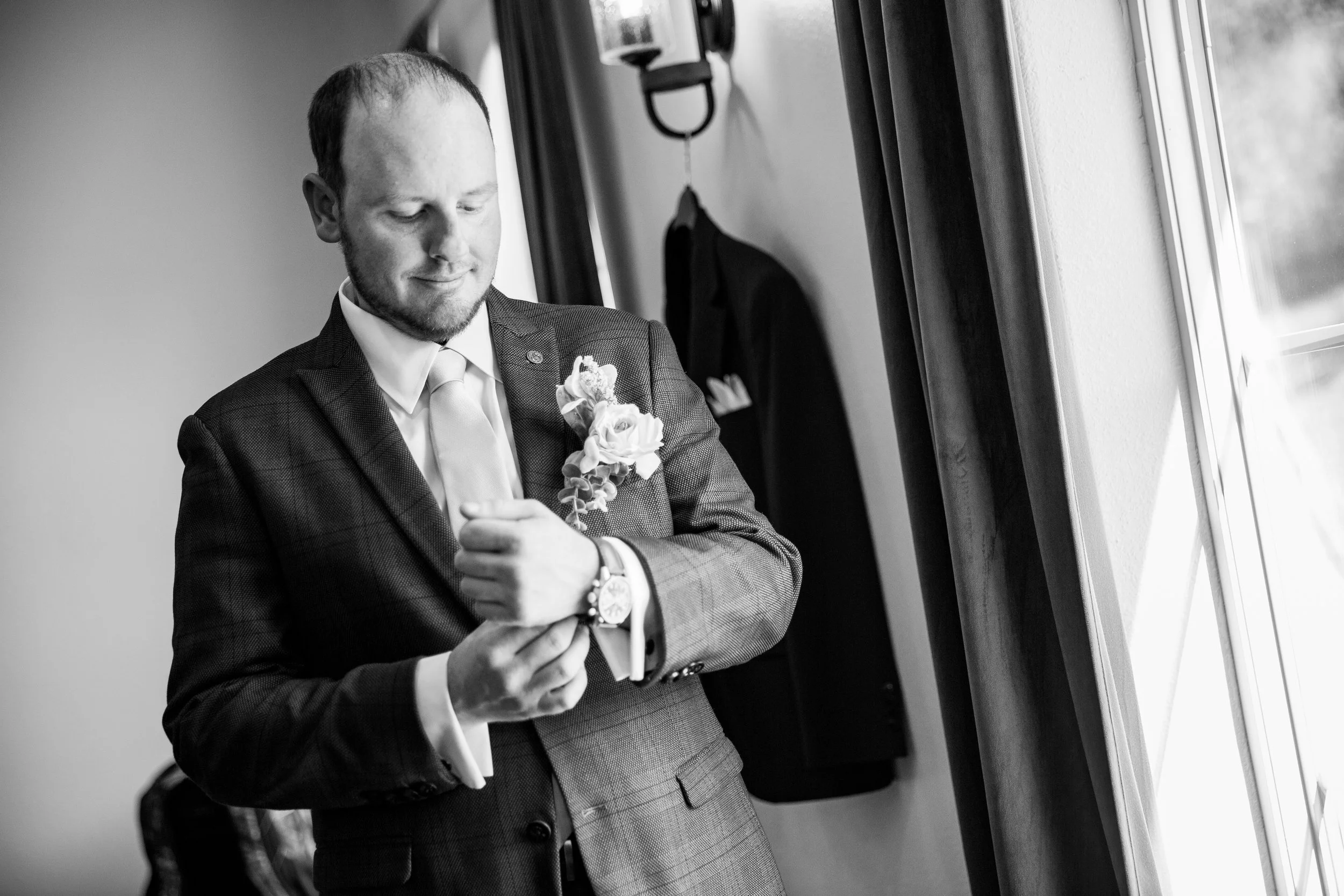 A man in a suit looking at a flower boutonniere on his lapel, standing near a window.