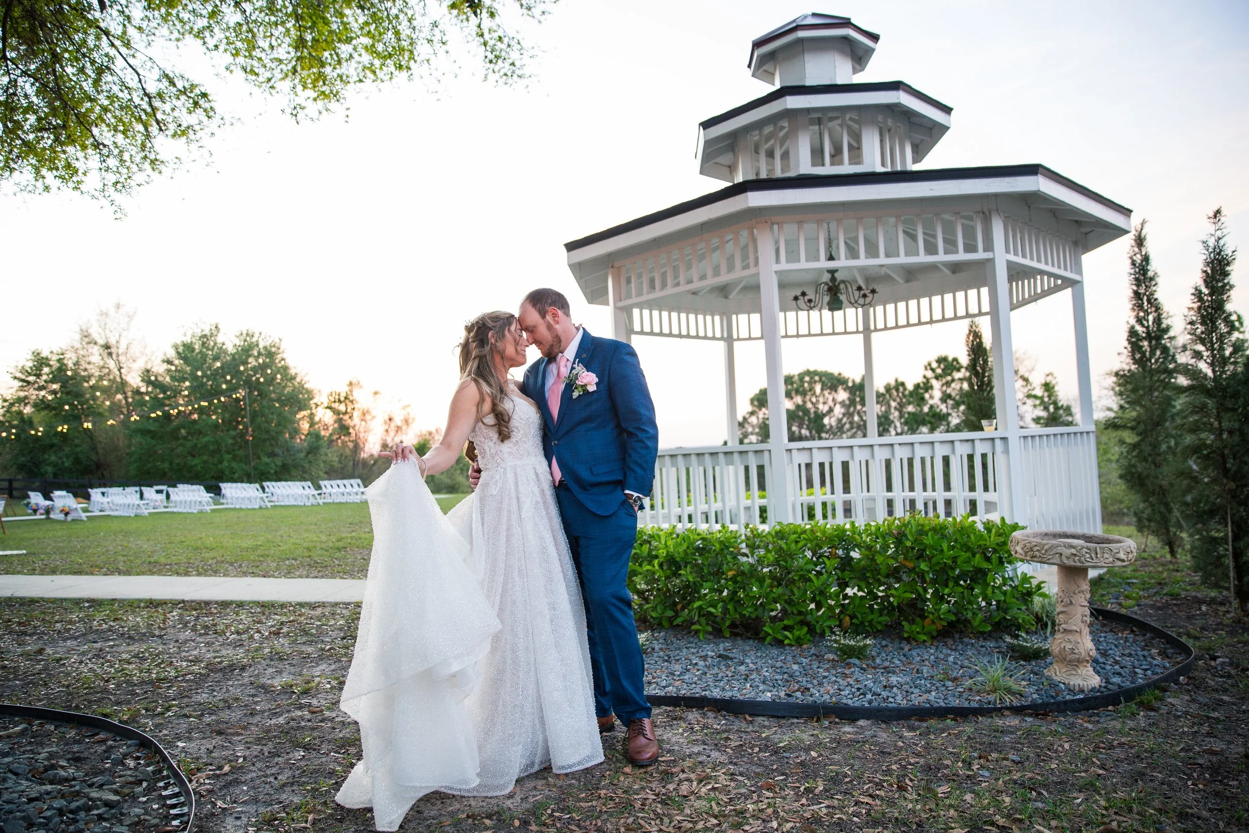 A bride and groom standing close together, touching foreheads, outdoors near a white gazebo, during sunset.