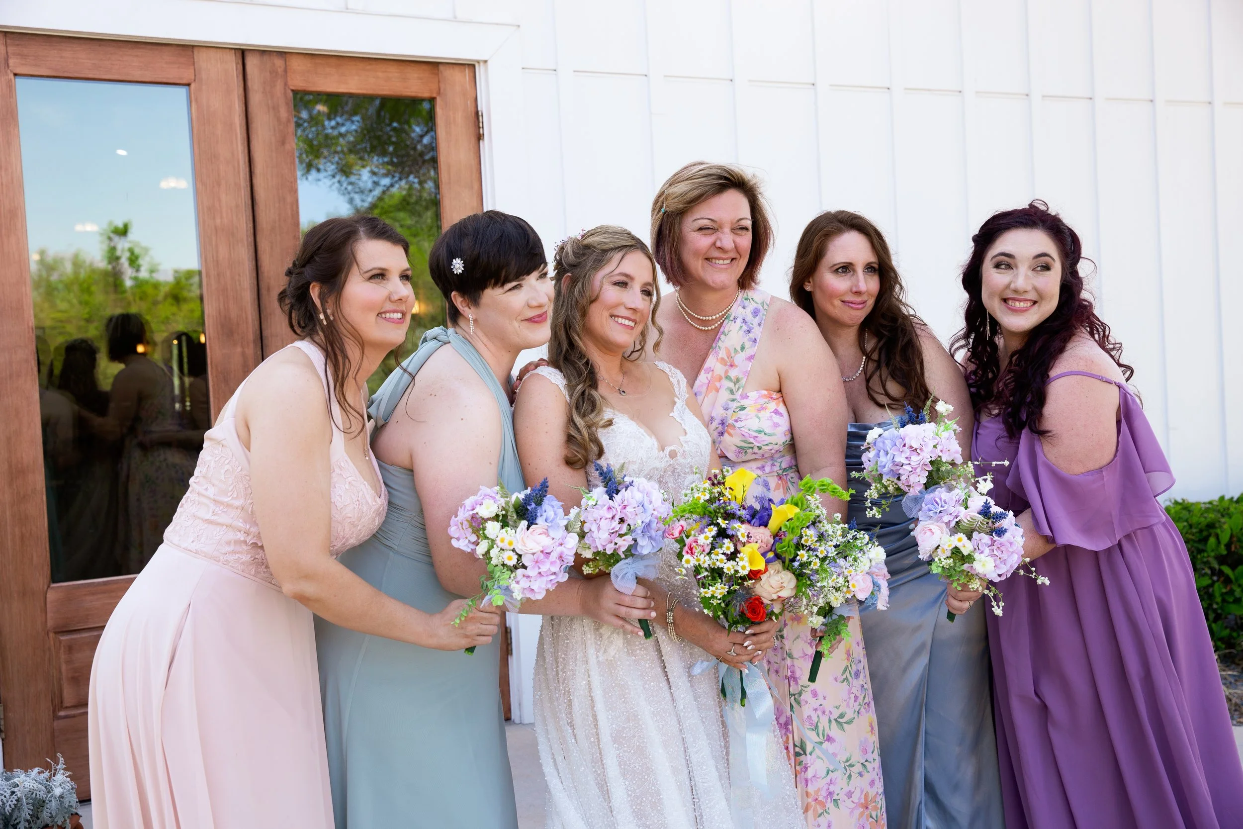 Group of women, including a bride in a white wedding dress, smiling and holding bouquets, standing outside near a white building with wooden doors.