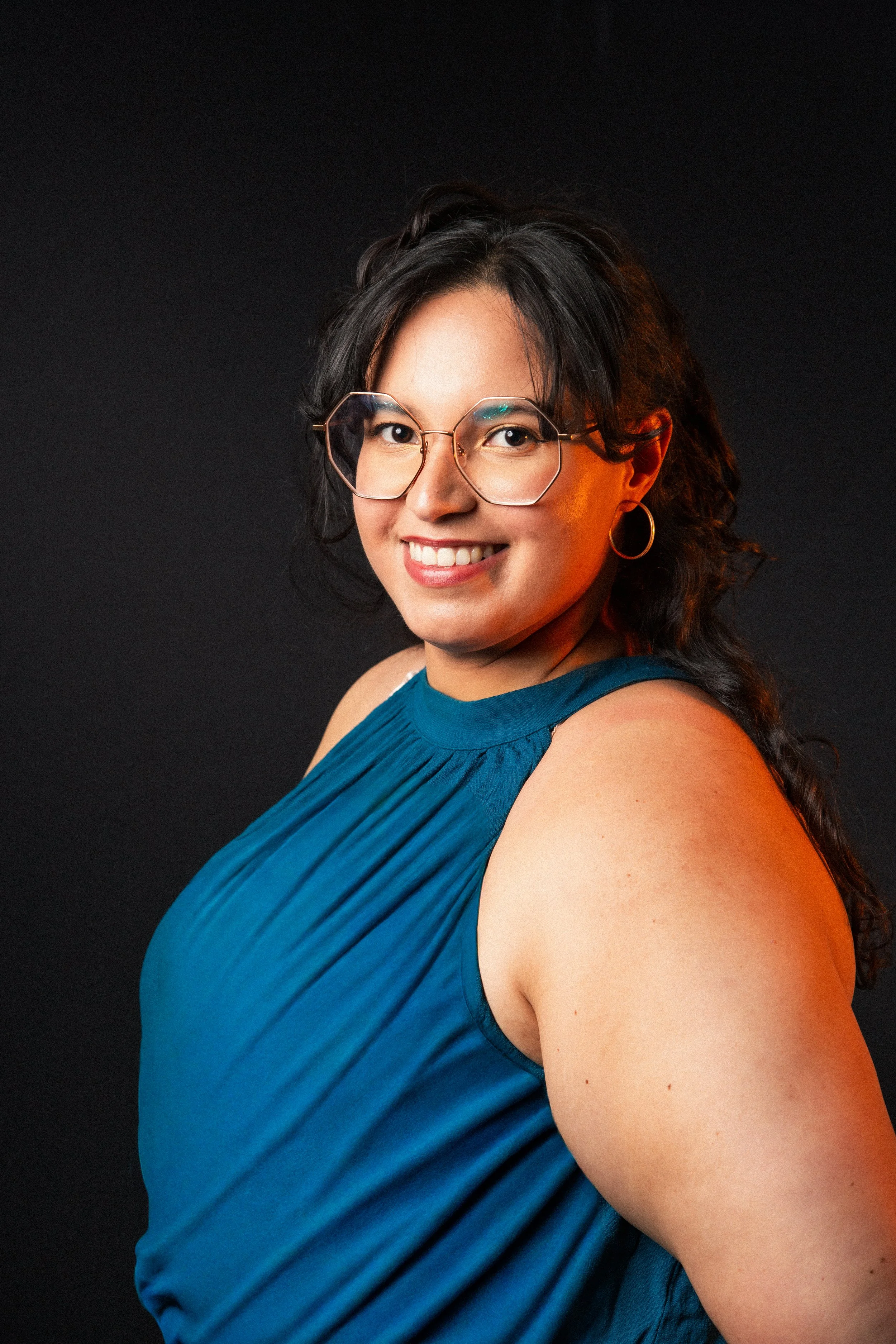 A woman with black hair, wearing glasses, hoop earrings, and a sleeveless blue top, smiling against a black background.
