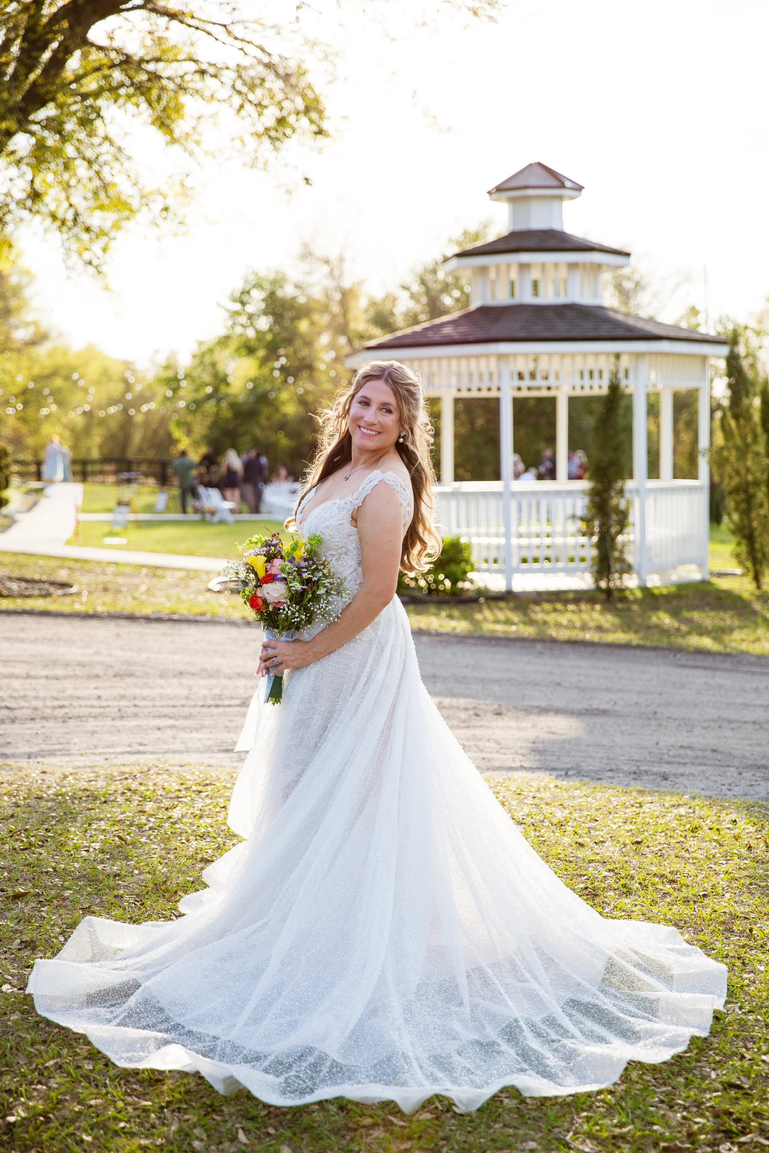 A bride in a white wedding gown holding a bouquet of flowers, smiling outdoors with a gazebo and guests in the background during sunset.