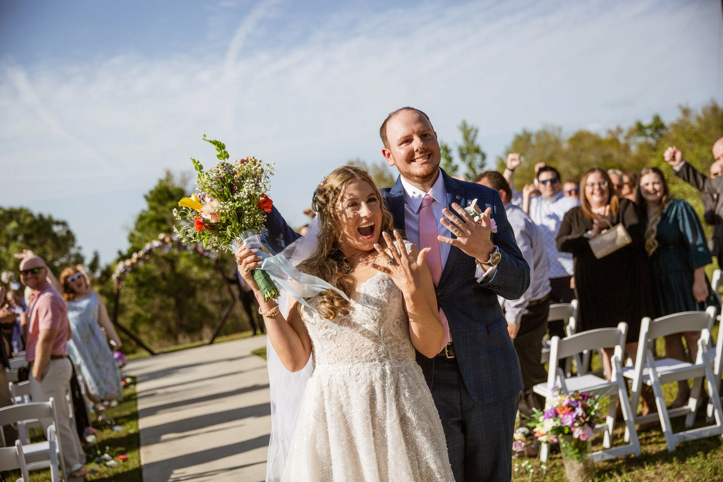 A newlywed couple celebrates after their wedding ceremony outdoors, with the bride holding a bouquet of flowers and both showing their wedding rings, while they are surrounded by friends and family cheering.
