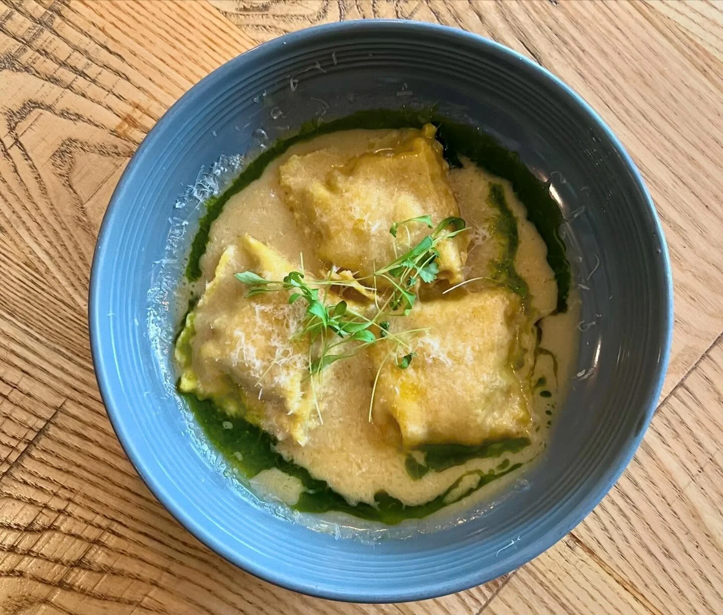 A blue bowl containing four pieces of ravioli topped with fresh herbs in a creamy sauce with a drizzle of green oil, placed on a wooden surface.