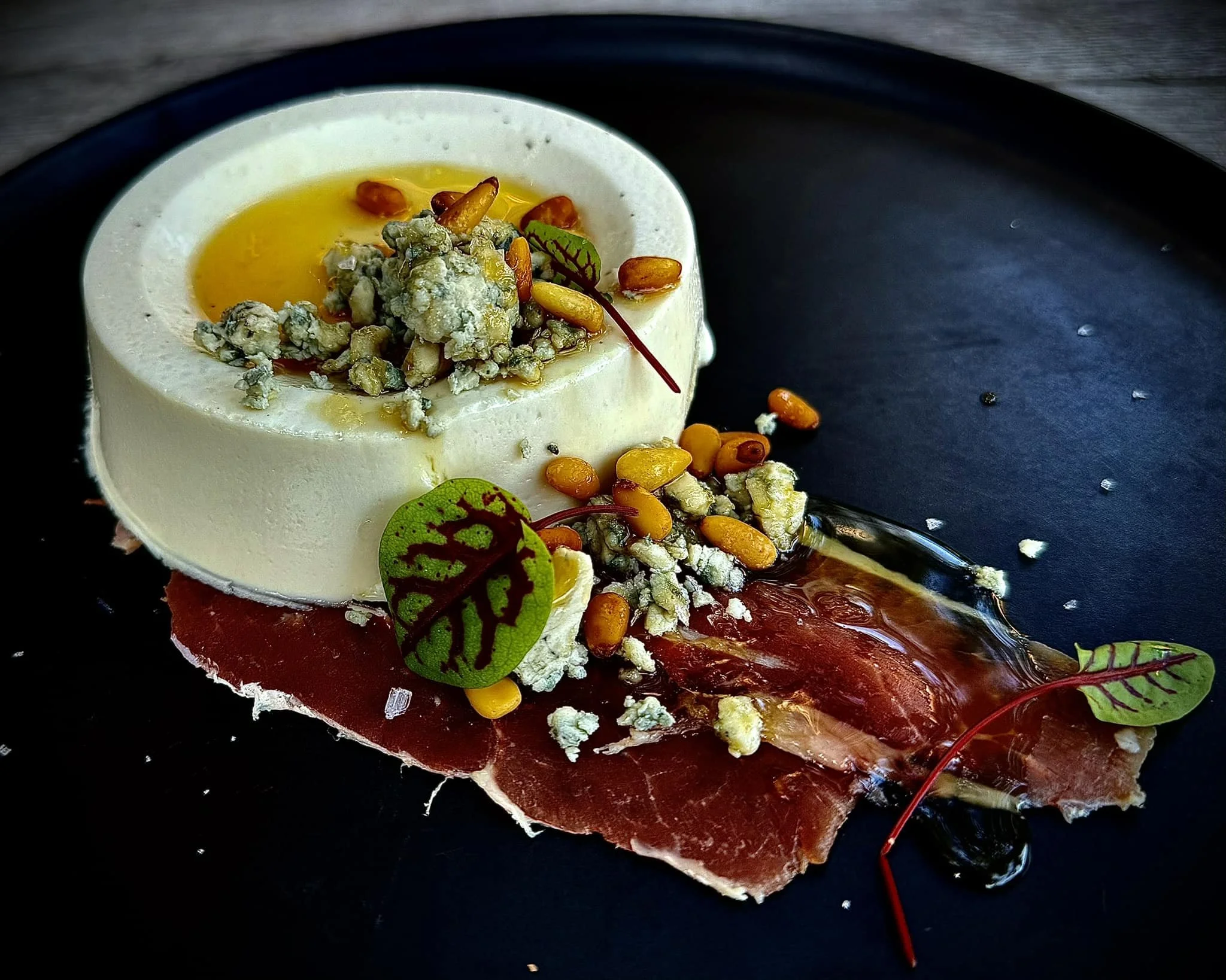 A dish featuring a round slice of creamy cheese topped with a raw egg yolk, blue cheese crumbles, pine nuts, and small leafy greens, served on a black plate with a slice of cured meat underneath and oil or balsamic drizzled on the side.