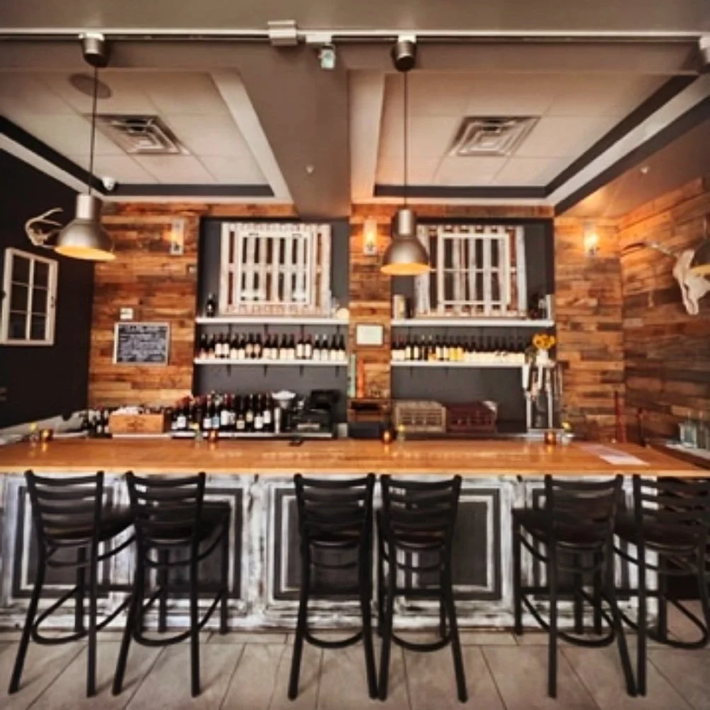 Bar with wooden counter, black chairs, wooden walls, and wine bottles on shelves behind the bar.