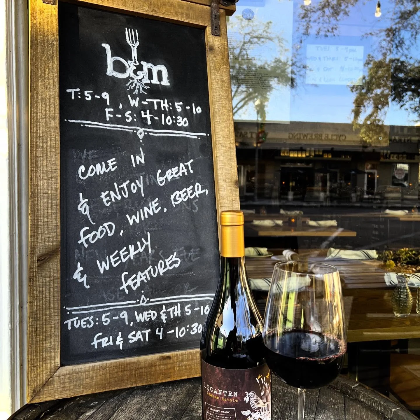 A chalkboard sign outside a restaurant with operating hours and a message inviting customers to enjoy food, wine, beer, and weekly features. In front of the sign, there is a bottle of red wine and a glass of red wine on a wooden surface.