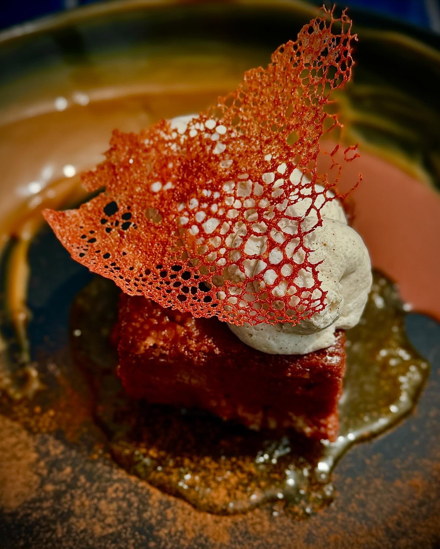 A gourmet dessert featuring a cube of red velvet cake topped with a swirl of light-colored frosting, garnished with a delicate, lacy red tuile, served on a textured dark plate with caramel and cinnamon dusting.