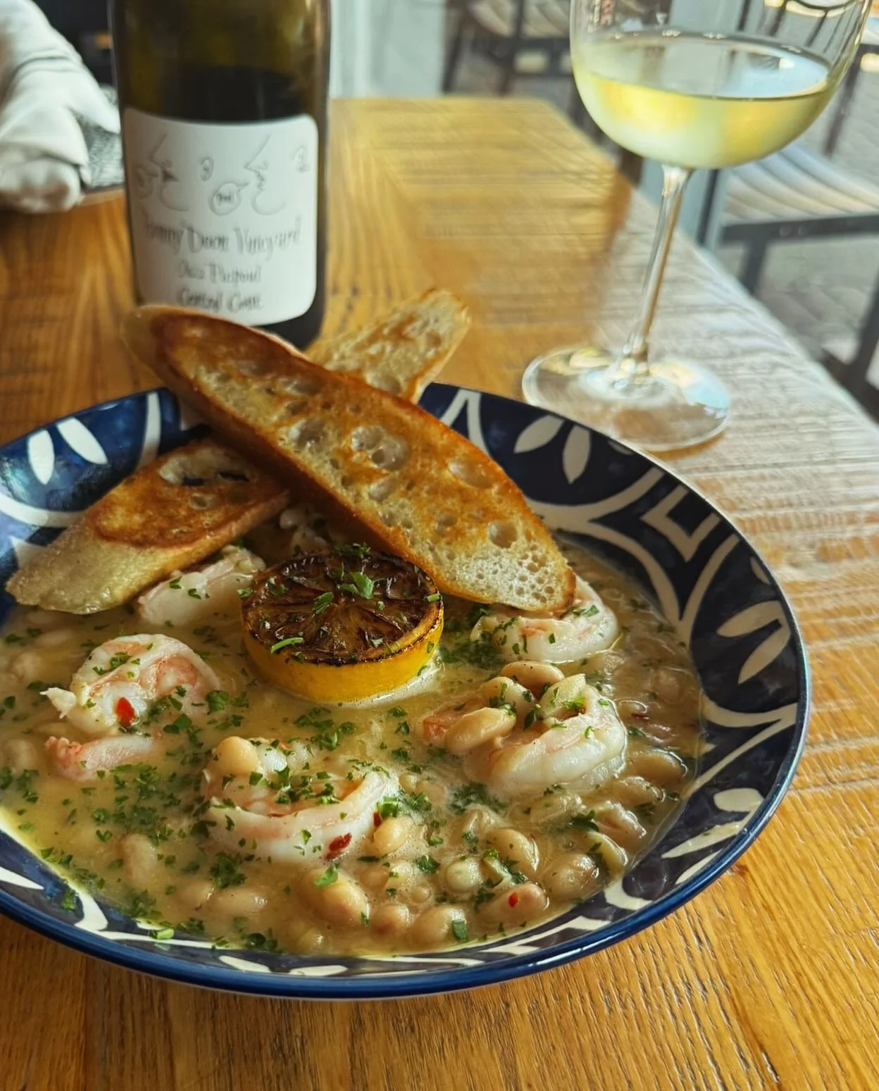 Shrimp and white bean stew garnished with chopped herbs, served with toasted bread slices and a grilled lemon half, with a glass of white wine and a bottle in the background.