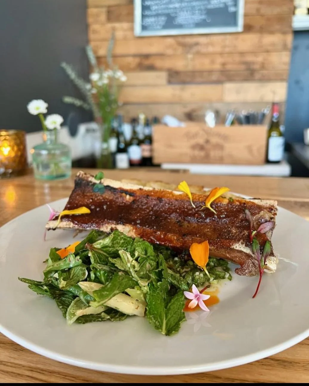 Plate with a large grilled rib, topped with barbecue sauce, garnished with edible flowers, and served with a side salad of greens and herbs on a white plate in a cozy restaurant setting.