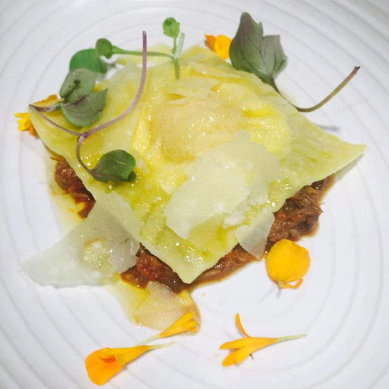 A plated dish with a crispy base topped with cheese, microgreens, and edible flowers, garnished with a yellow sauce.