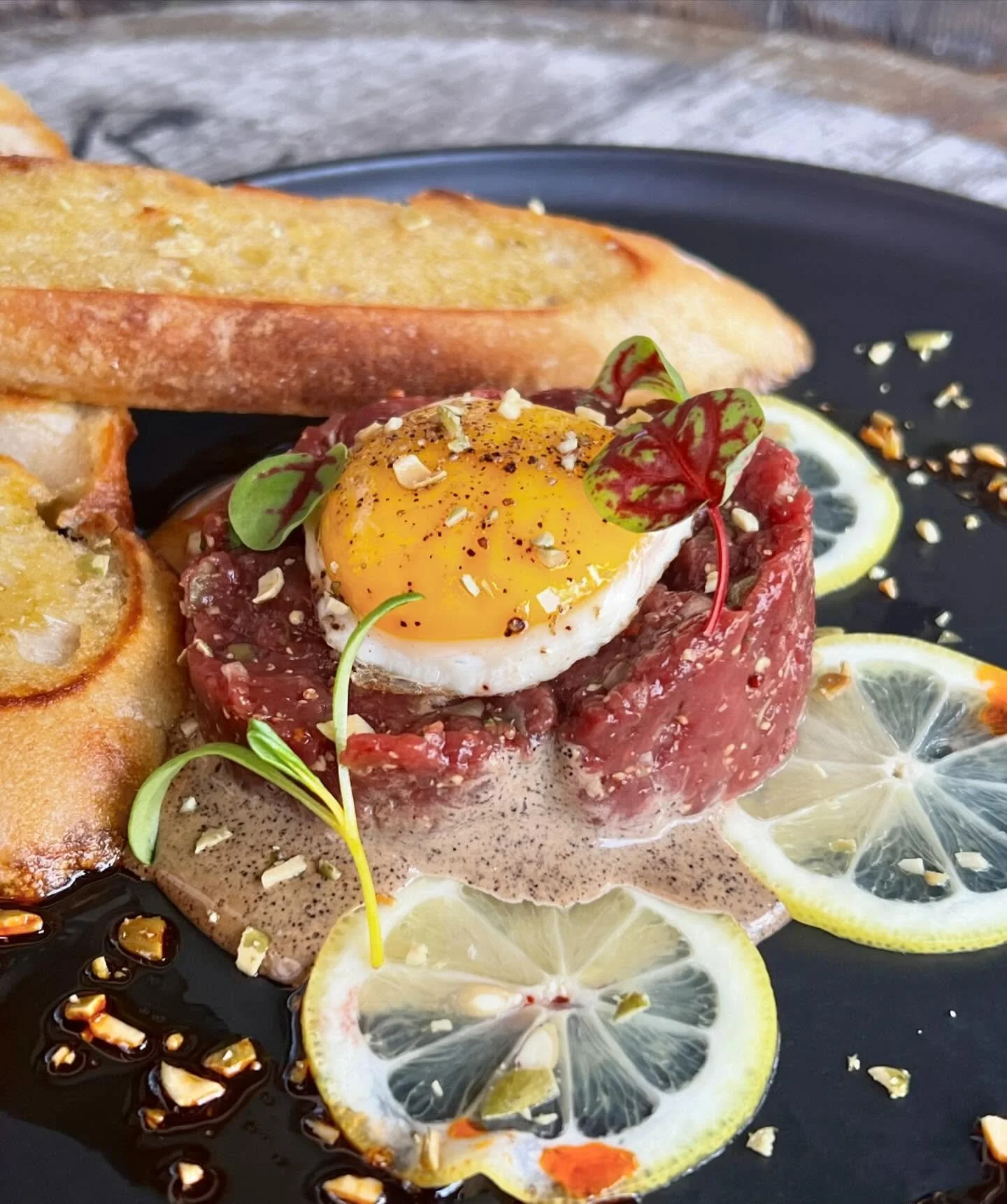 Steak tartare topped with a poached egg, garnished with microgreens, served with lemon slices, toasted bread, and sauce on a black plate.