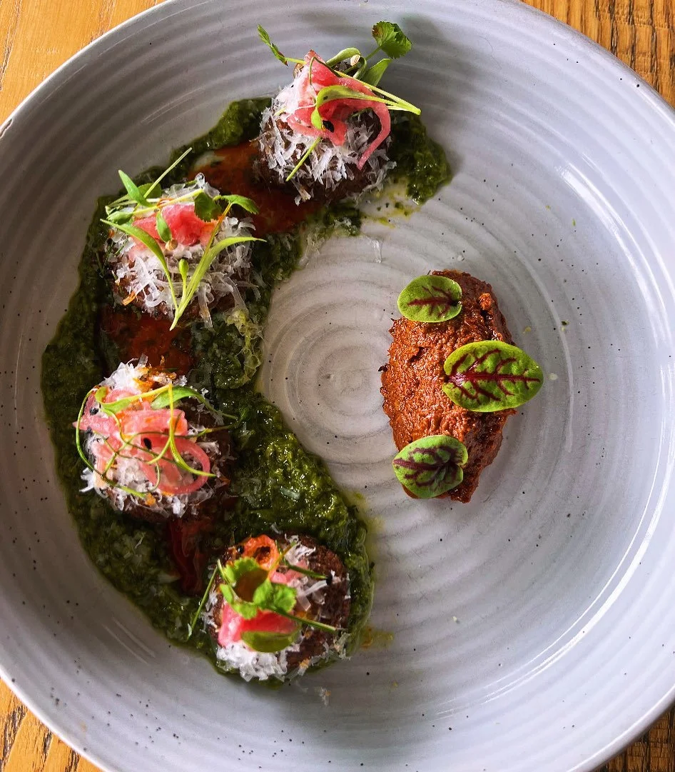 Gourmet dish with four meat stuffed balls garnished with microgreens and grated cheese, drizzled with dark sauce, next to a rectangular fried patty topped with small green leaves, all on a large grey plate.