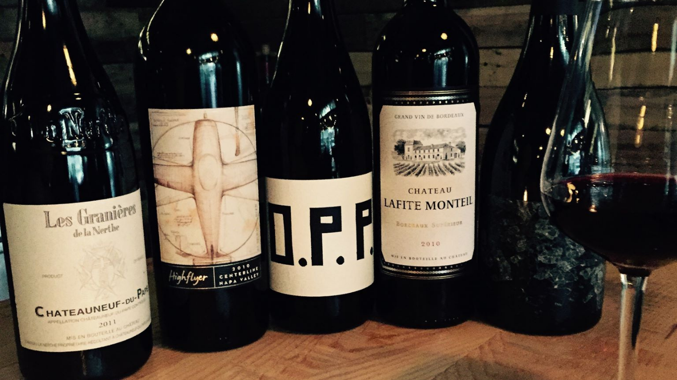 Five wine bottles lined up on a wooden surface with a glass of red wine next to them.