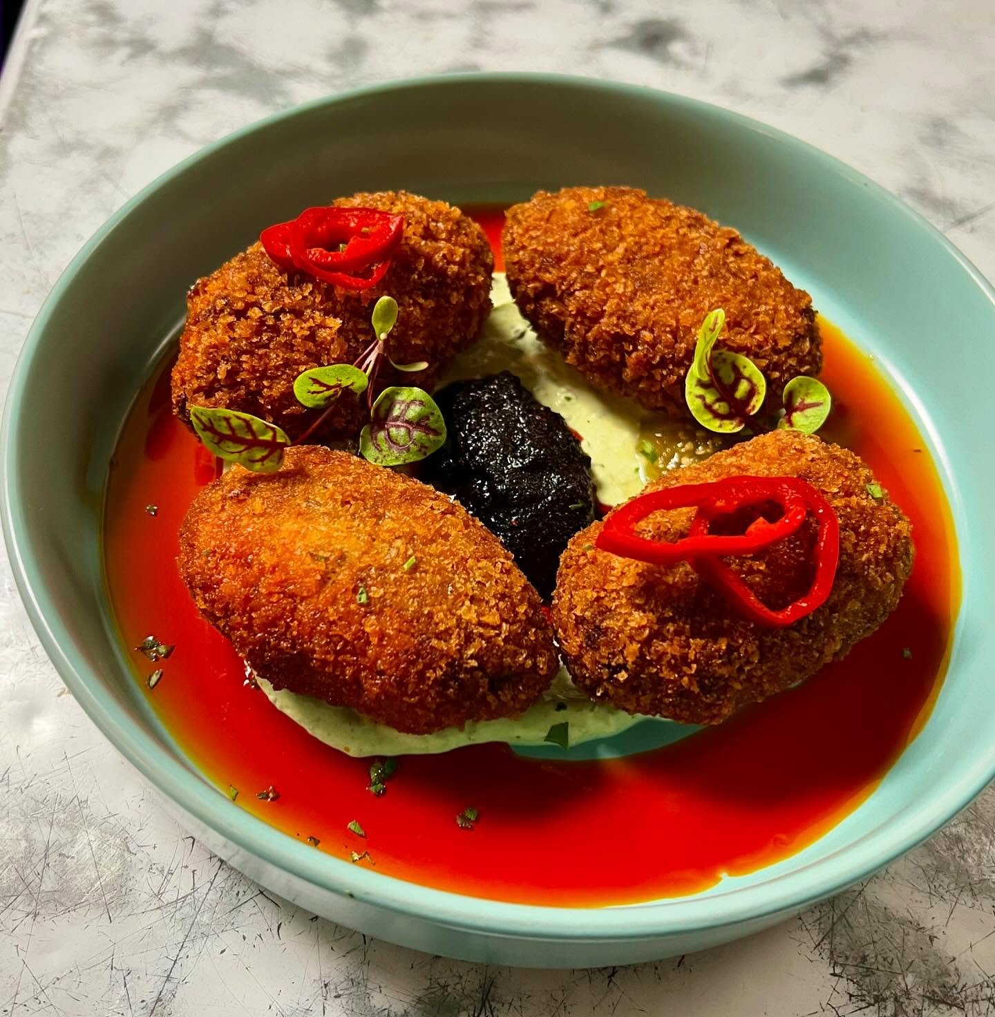 Fried croquettes with red chili slices, microgreens, and black sauce on a bed of green sauce, served in a blue bowl with red sauce around the edge.