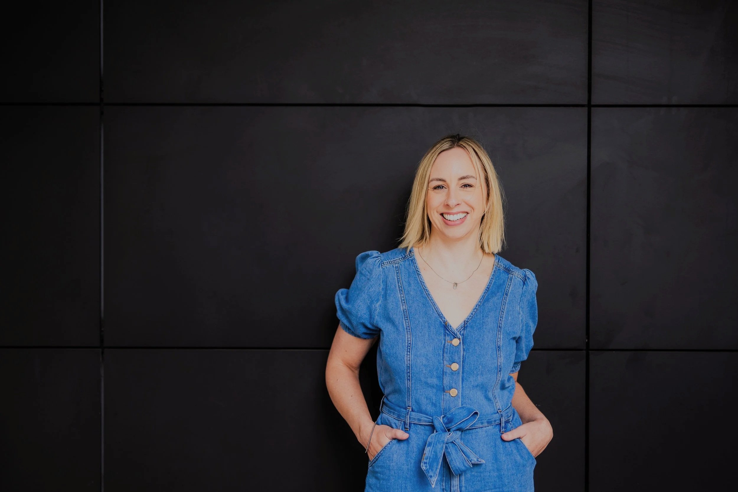 A woman with blonde hair, wearing a blue denim jumpsuit with a tied belt, smiling and standing against a black wall.