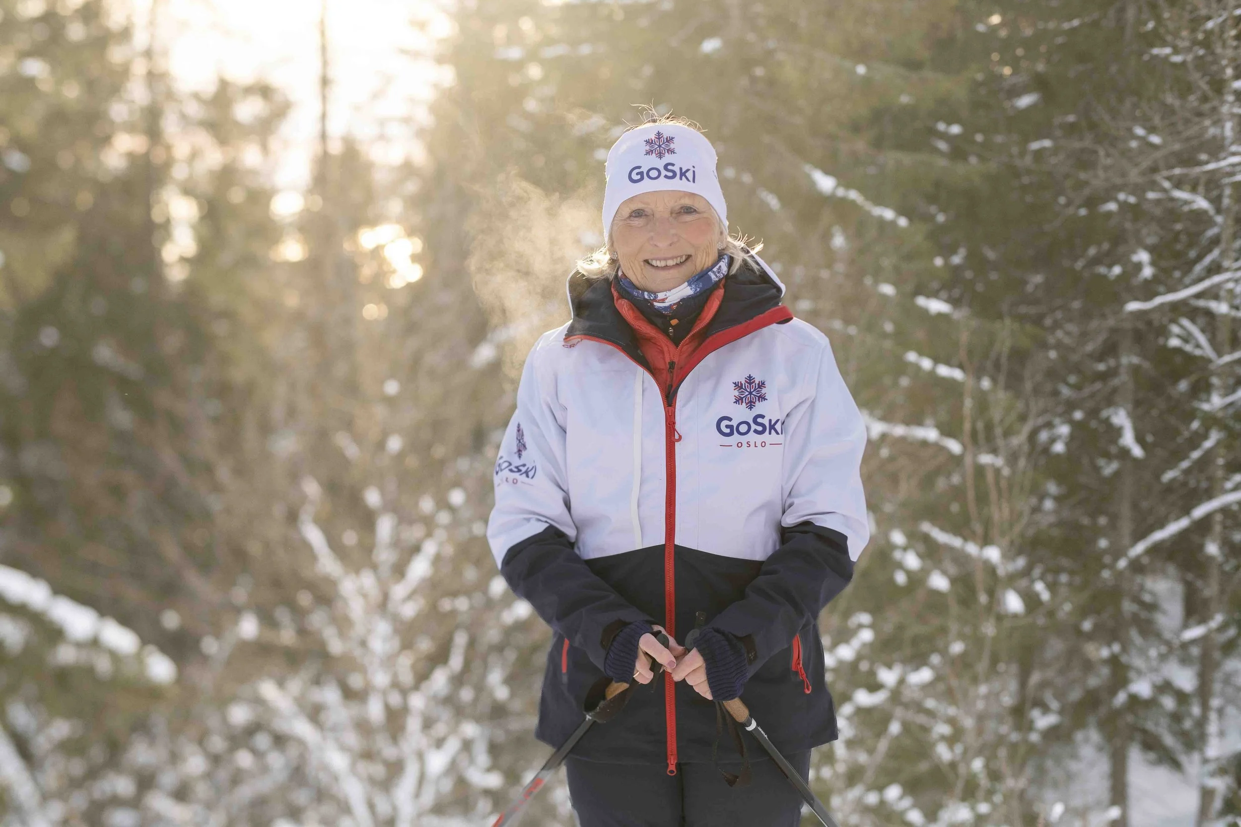 En eldre kvinne i vintertøy og lue som smiler, står i snøfylt skog med skiutstyr i hendene. Foto: Benjamin Haug-Toklum