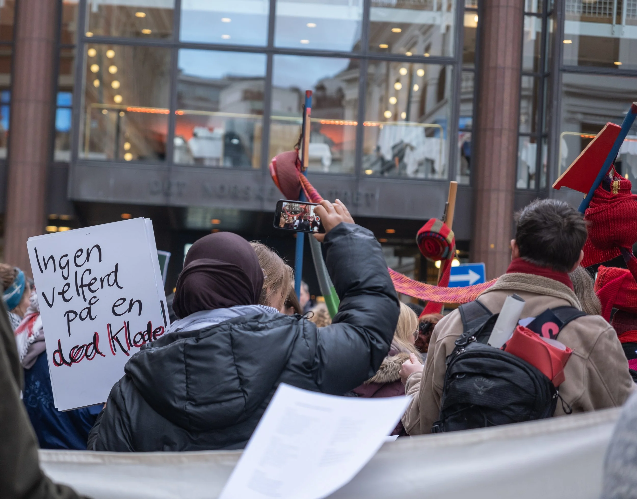 Folk som deltar i en demonstrasjon, noen holder opp bannere og flagg, foran en bygning med glassfasade. Foto: Benjamin Haug-Toklum