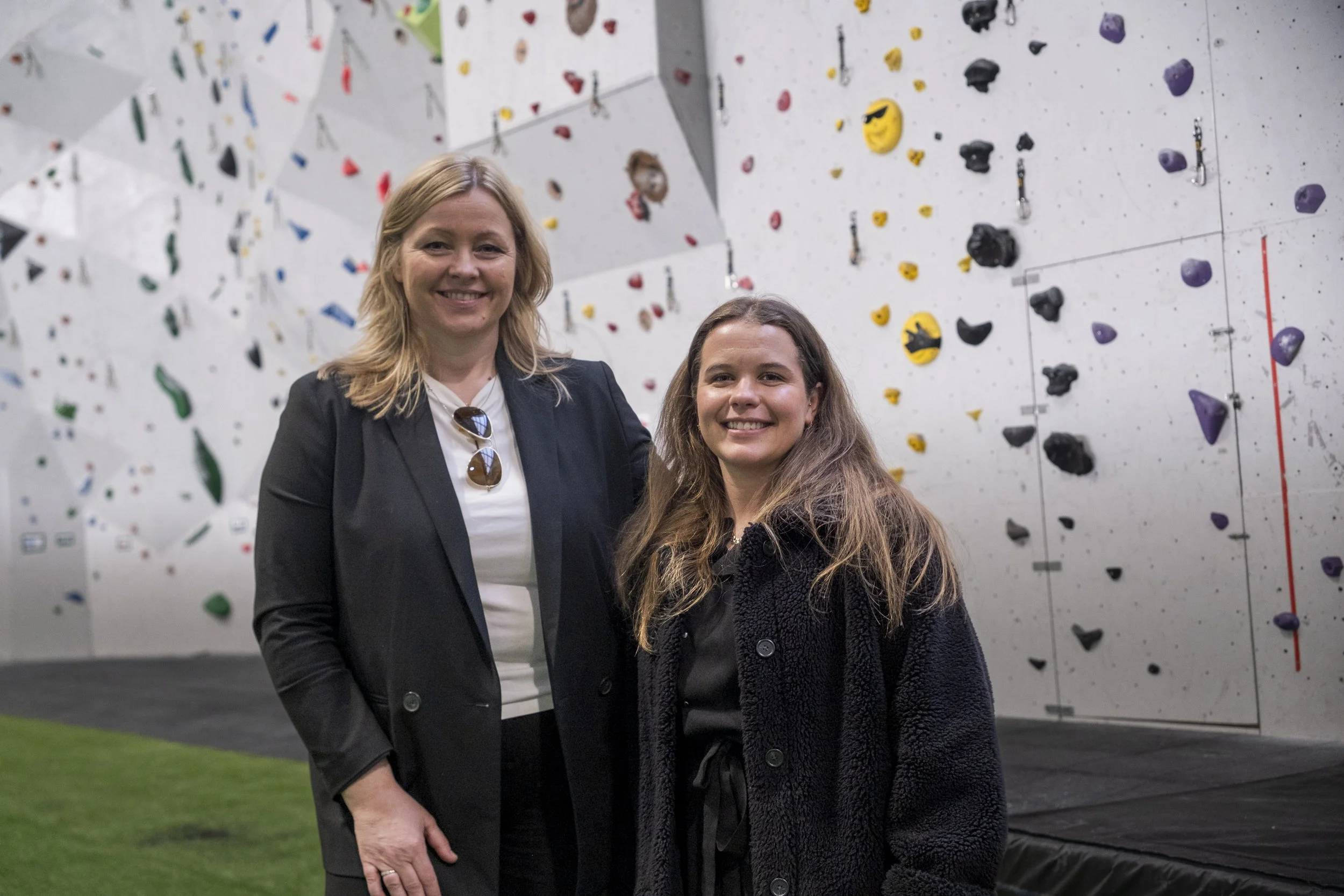 To kvinner står foran en innendørs klatrevegg, smilende. Klatreveggen har forskjellige fargede håndtak og skruebikter. Foto: Benjamin Haug-Toklum