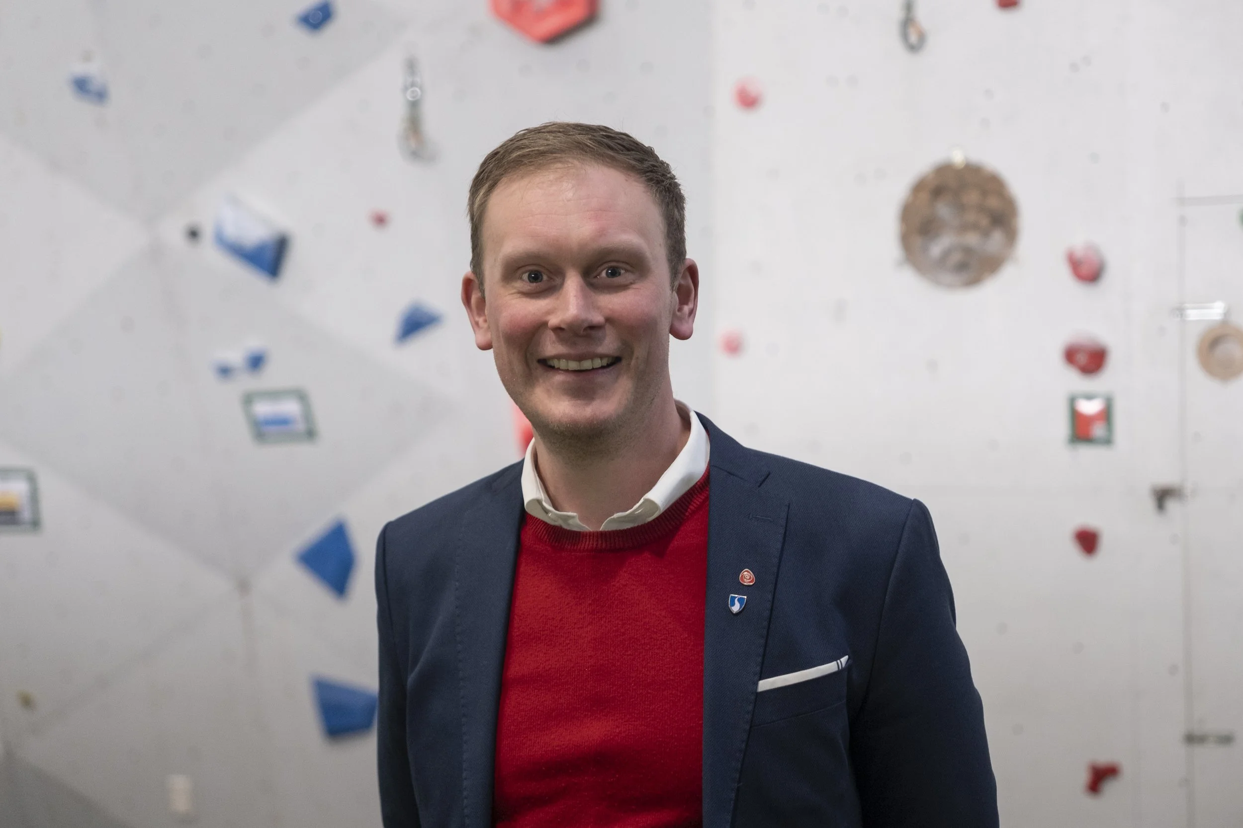 Sogndal ordfører Stig Ove Ølmheim (AP) med mørkeblå dress, hvit skjorte og rød genser smiler foran en innendørs klatrevegg. Foto: Benjamin Haug-Toklum