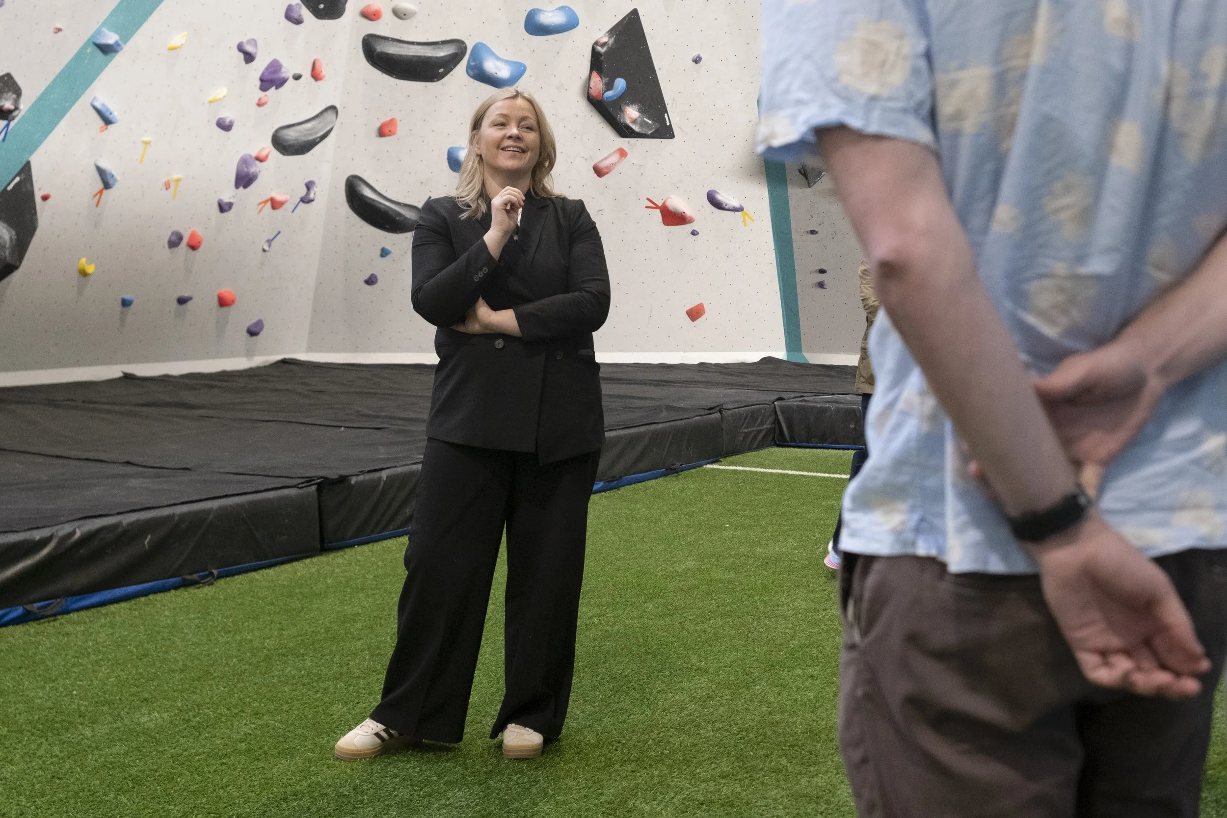 En kvinne, Kommunal- og distriktsminister
Kjersti Stenseng (Ap), i svart dress smiler mens hun snakker til en mann i en innendørs klatrehall, med fargede klatregrep på veggen bak henne. Foto: Benjamin Haug-Toklum