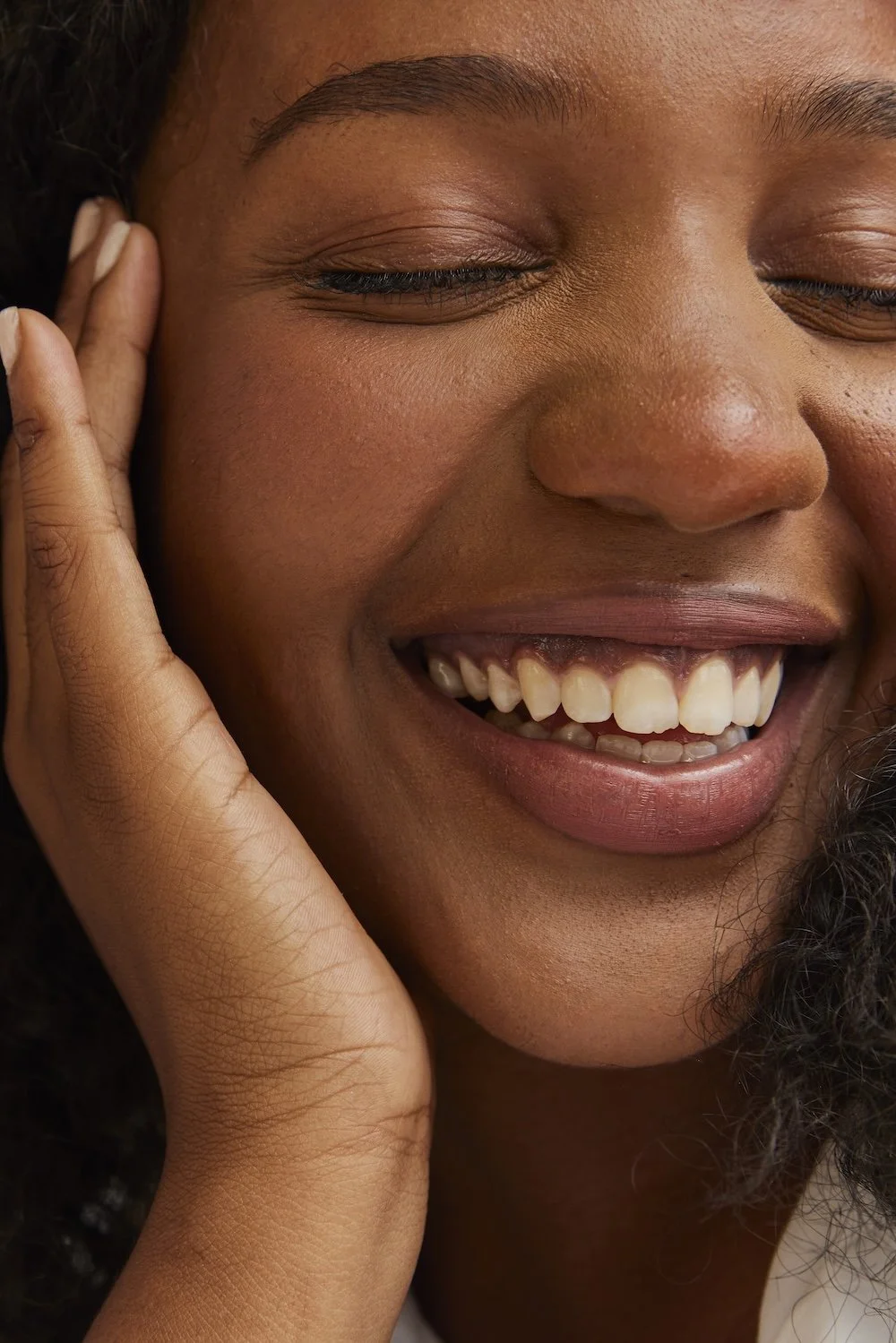 Close-up of a smiling Black woman with eyes gently closed, showing her teeth, with a hand touching her face.