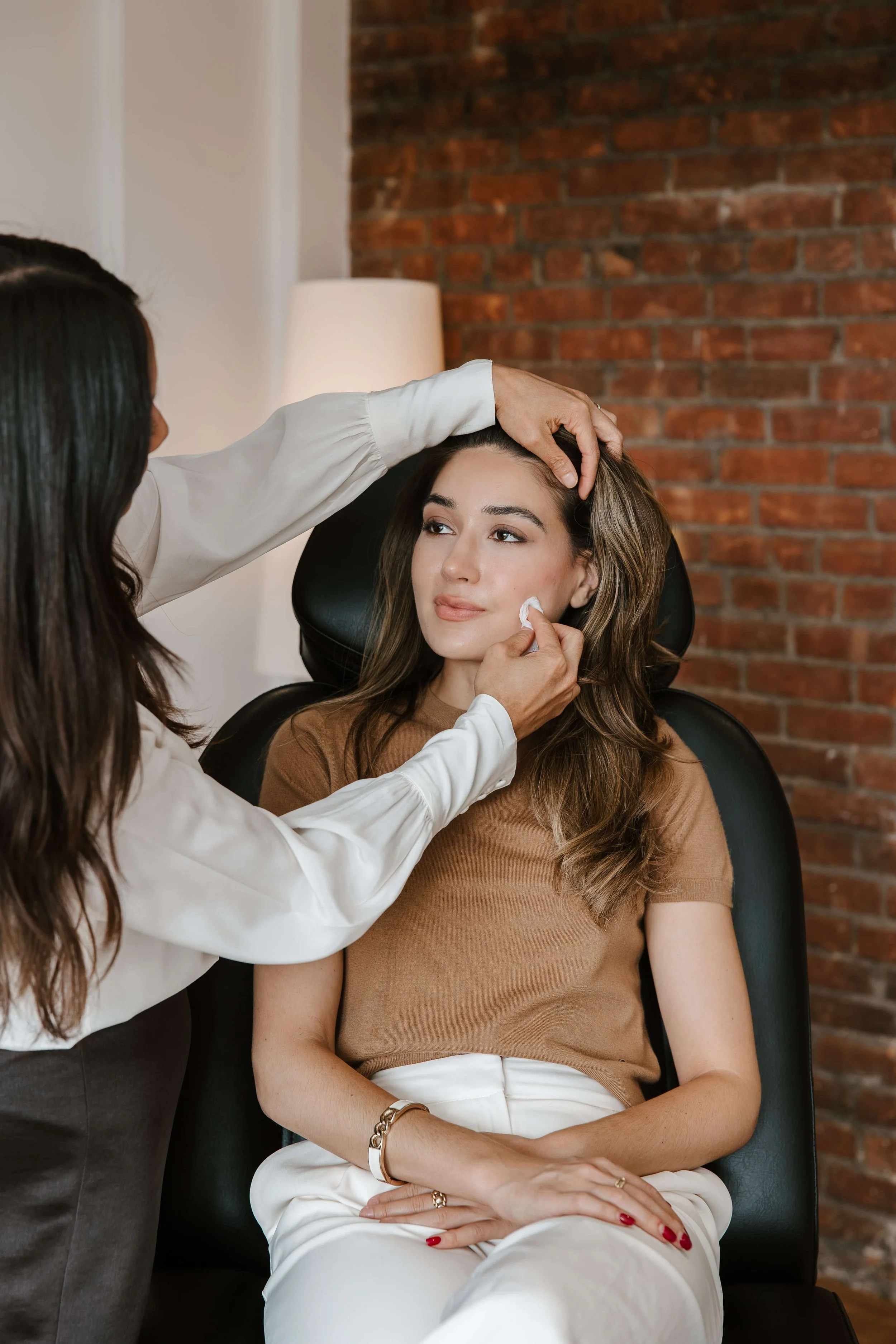 A woman with long brown hair sitting in a chair is getting her face groomed by a beautician, with a brick wall in the background.