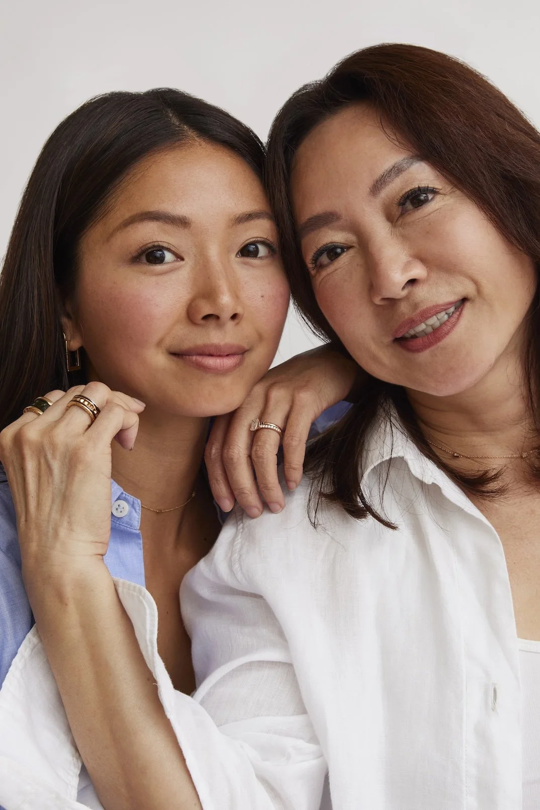 Two women close together, smiling, with one resting her hand on the other's shoulder, wearing necklaces and rings.