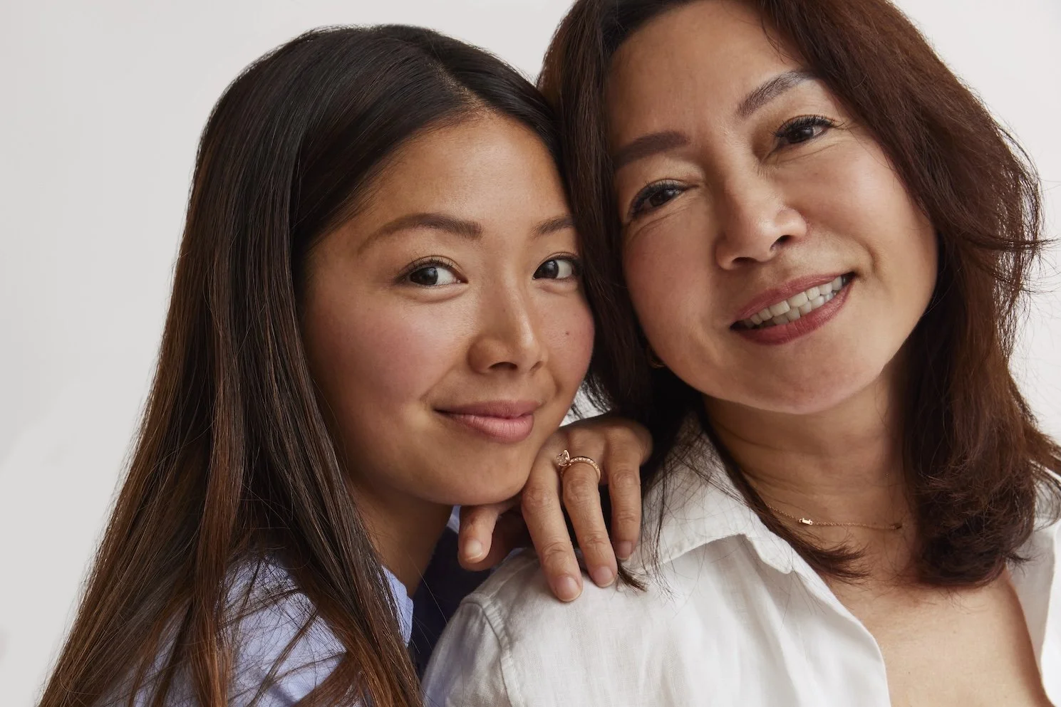 Close-up of two women smiling, one with long dark hair and the other with shoulder-length red hair, in a white shirt, against a plain background.