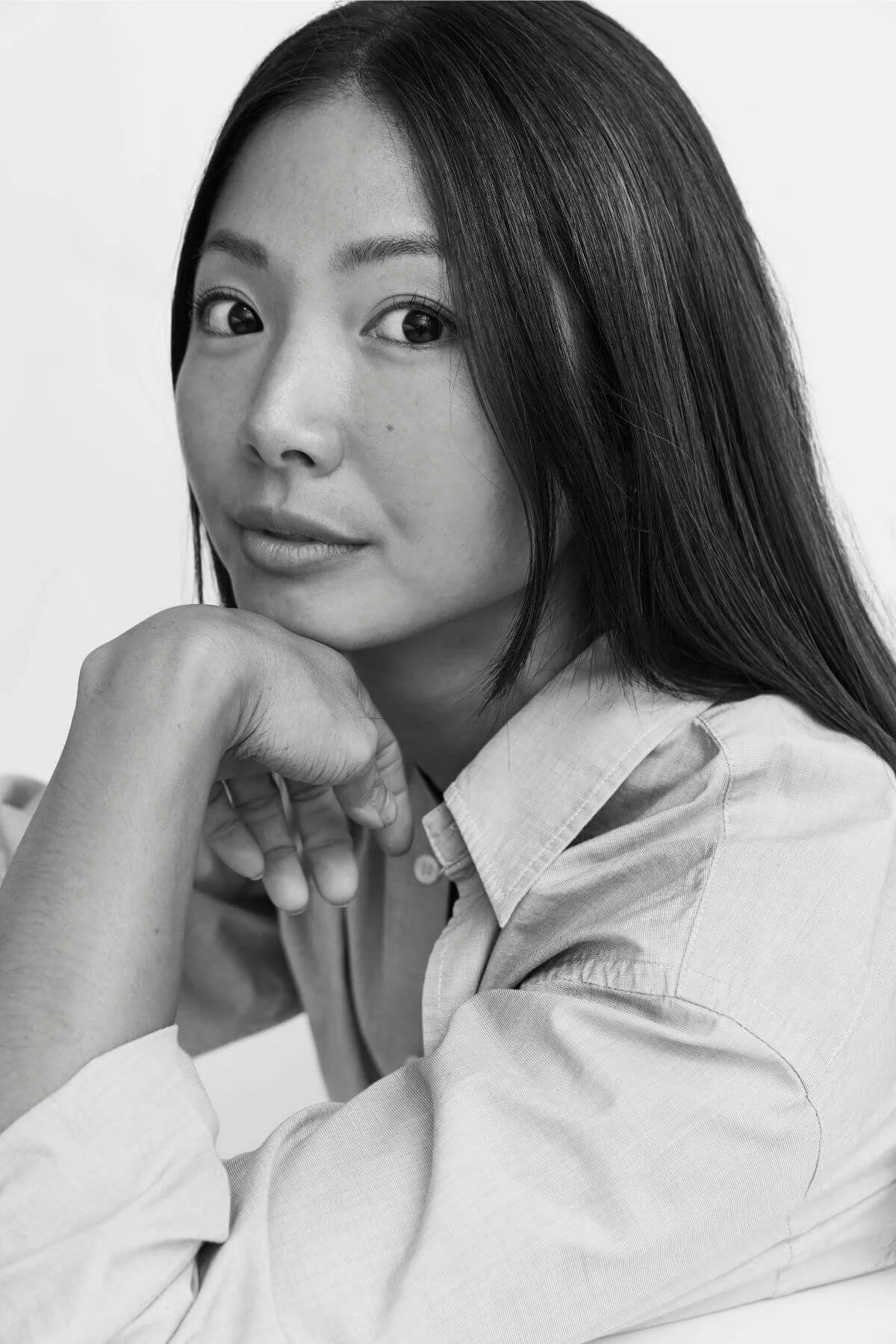 Black and white portrait of a woman with long dark hair, resting her chin on her hand, wearing a light-colored shirt.