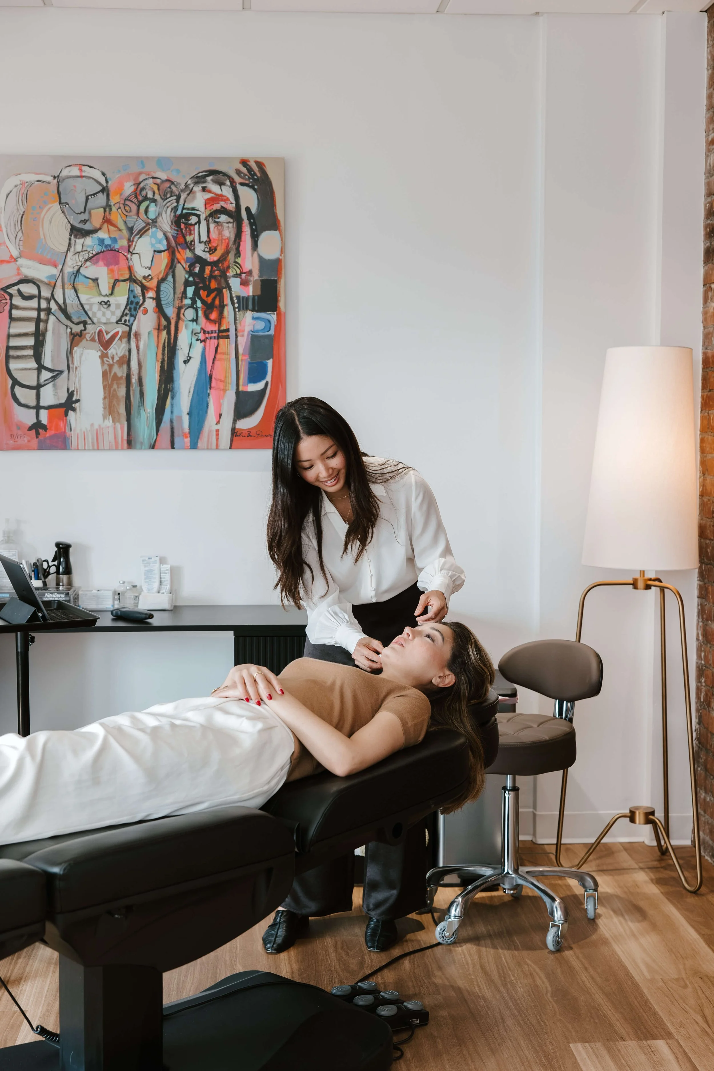 A woman lying on an examination table receiving a facial treatment from a skincare professional in a modern spa or clinic setting, with a colorful abstract painting on the wall and a tall white floor lamp nearby.