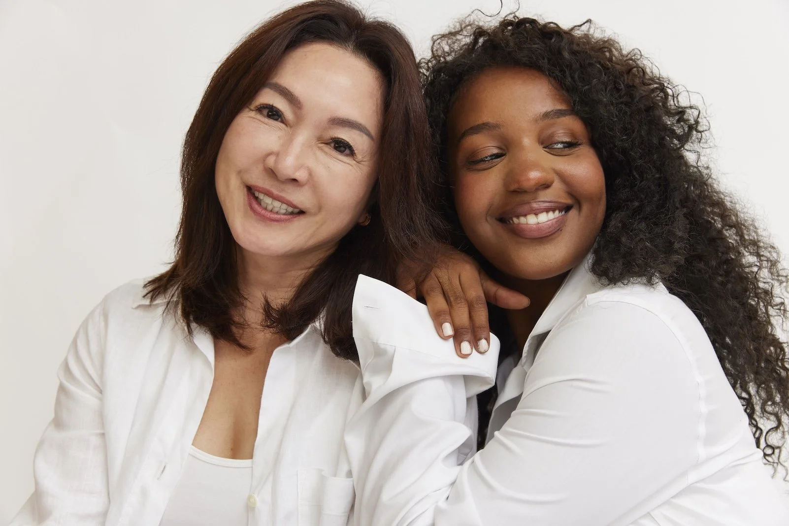 Two women smiling and embracing, one with straight hair and the other with curly hair, both wearing white shirts, against a plain white background.