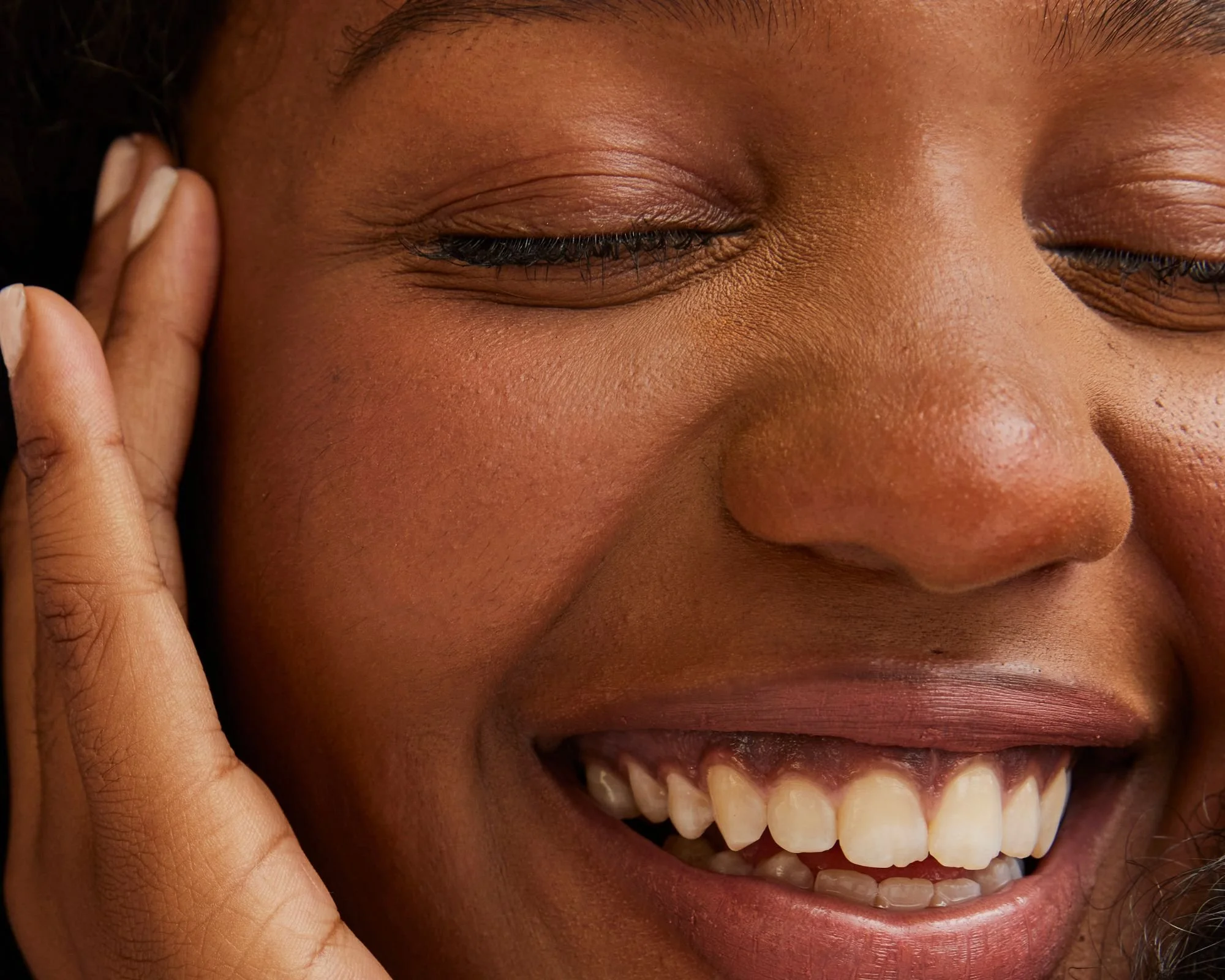 Close-up of a smiling woman's face with her eyes closed, showing white teeth, and a hand gently touching her cheek.