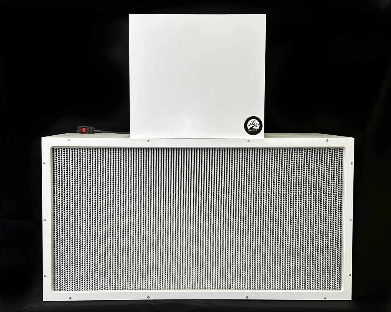 A white electronic device with a small white box on top and a large rectangular speaker with a grid front panel. The device has a black control knob and a power cord plugged into the back.