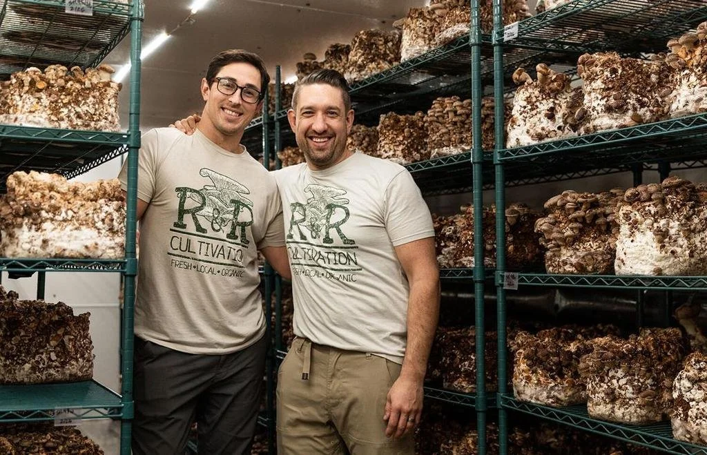 Two men standing in front of shelves filled with large blocks of cultivated mushrooms, smiling and wearing matching T-shirts with a mushroom logo and text.