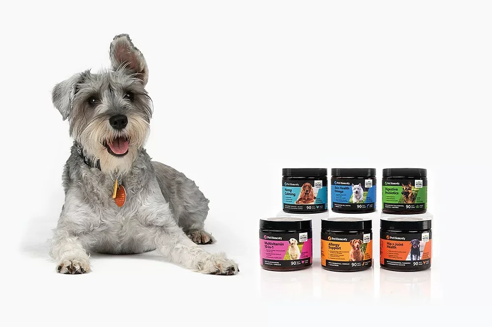A smiling dog sitting next to six containers of pet supplement products on a white background.