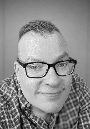 A man with short hair, glasses, and a checkered shirt smiling at the camera.