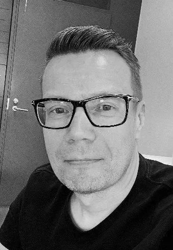 Black-and-white photo of a man with glasses, short hair, and slight beard, looking at the camera in an indoor setting.