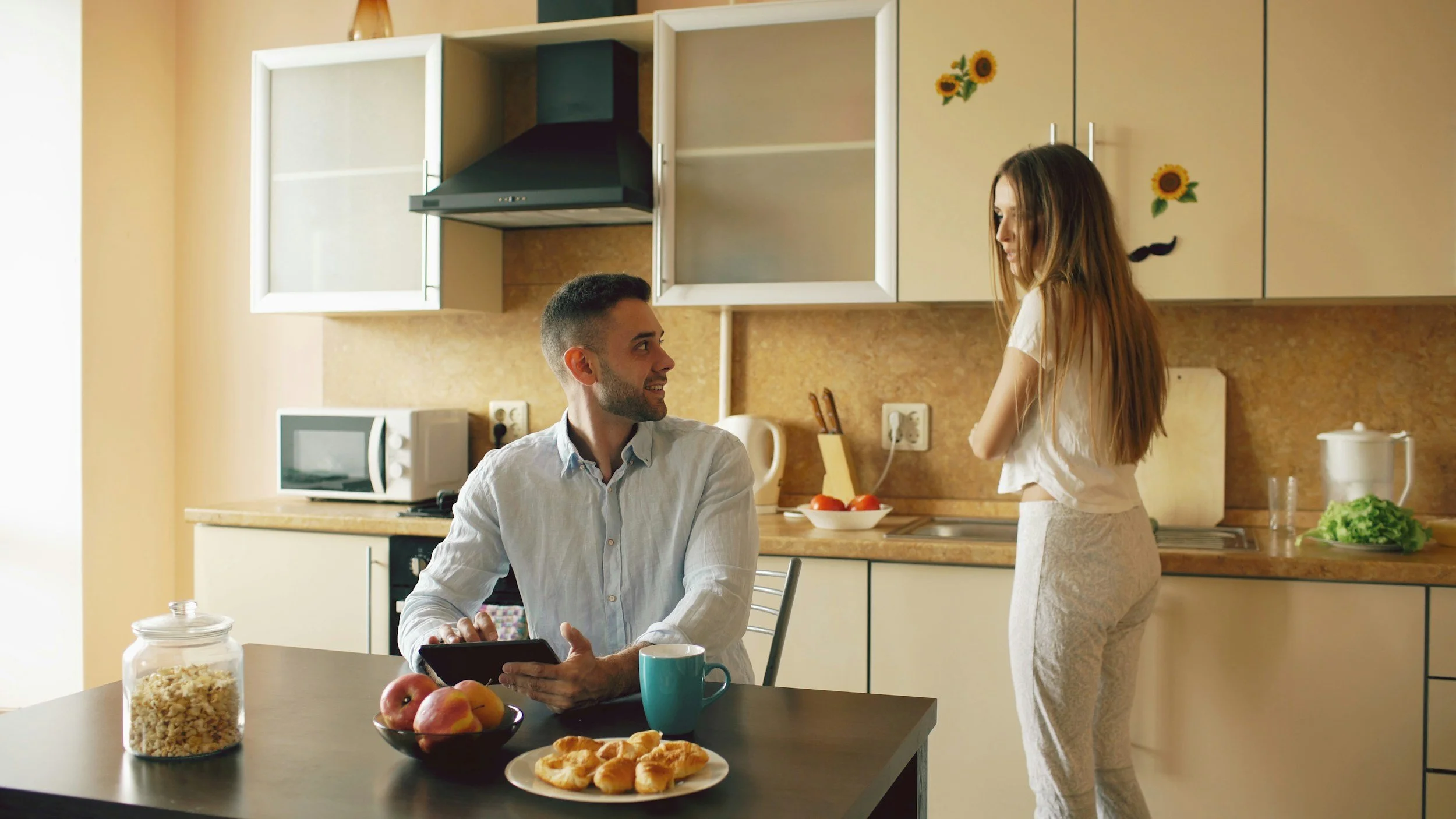 A man sitting at a kitchen table with a tablet, a bowl of apples, a mug, and croissants, while a woman stands nearby in a kitchen with cabinets, a microwave, and a countertop with a bowl of tomatoes and a cutting board.