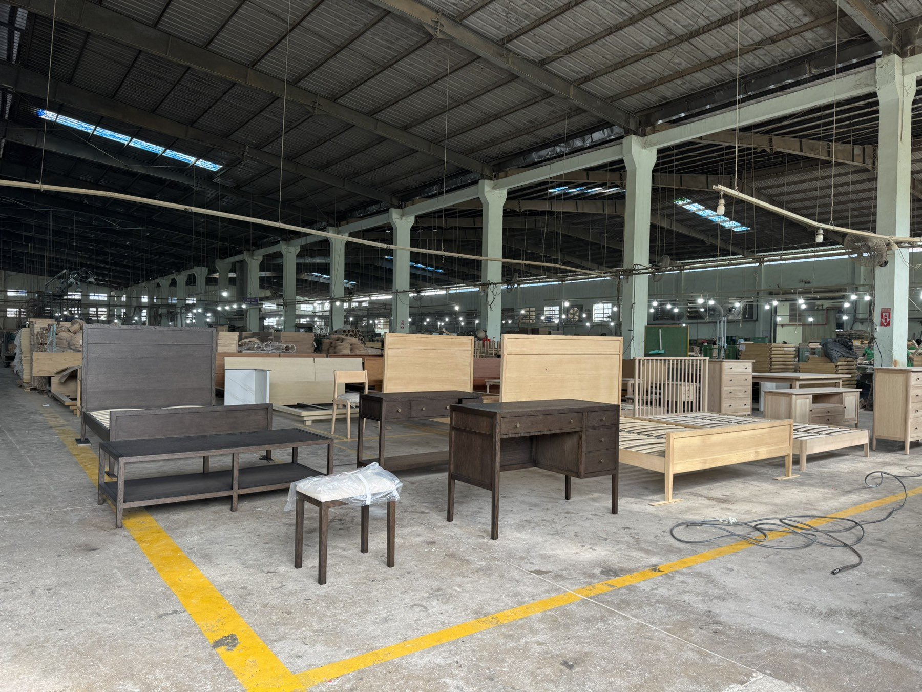 Warehouse with various unfinished wooden furniture pieces including beds, tables, and chairs, within a spacious industrial setting.