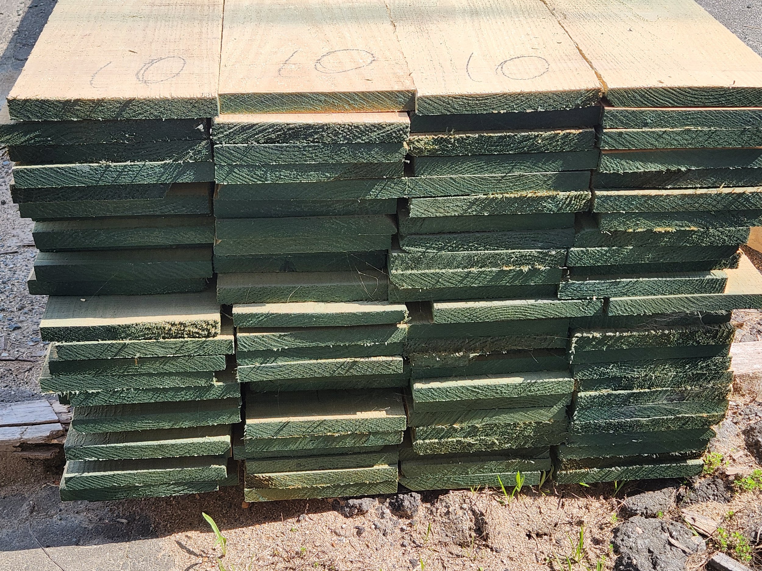 Stacked pieces of cut lumber with green paint or stain, placed on dirt ground outdoors.