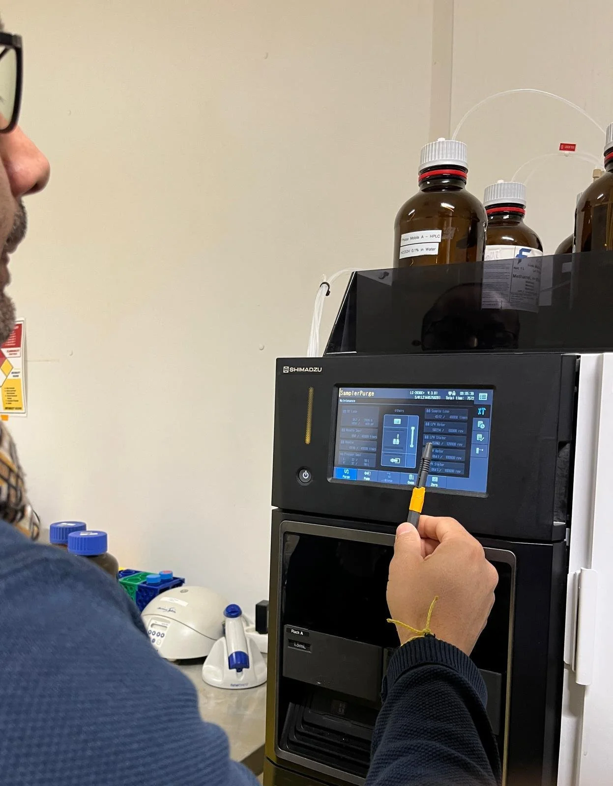 A scientist wearing gloves and a face shield holding a plastic tray containing six petri dishes with purple and patterned bacterial cultures in a laboratory.