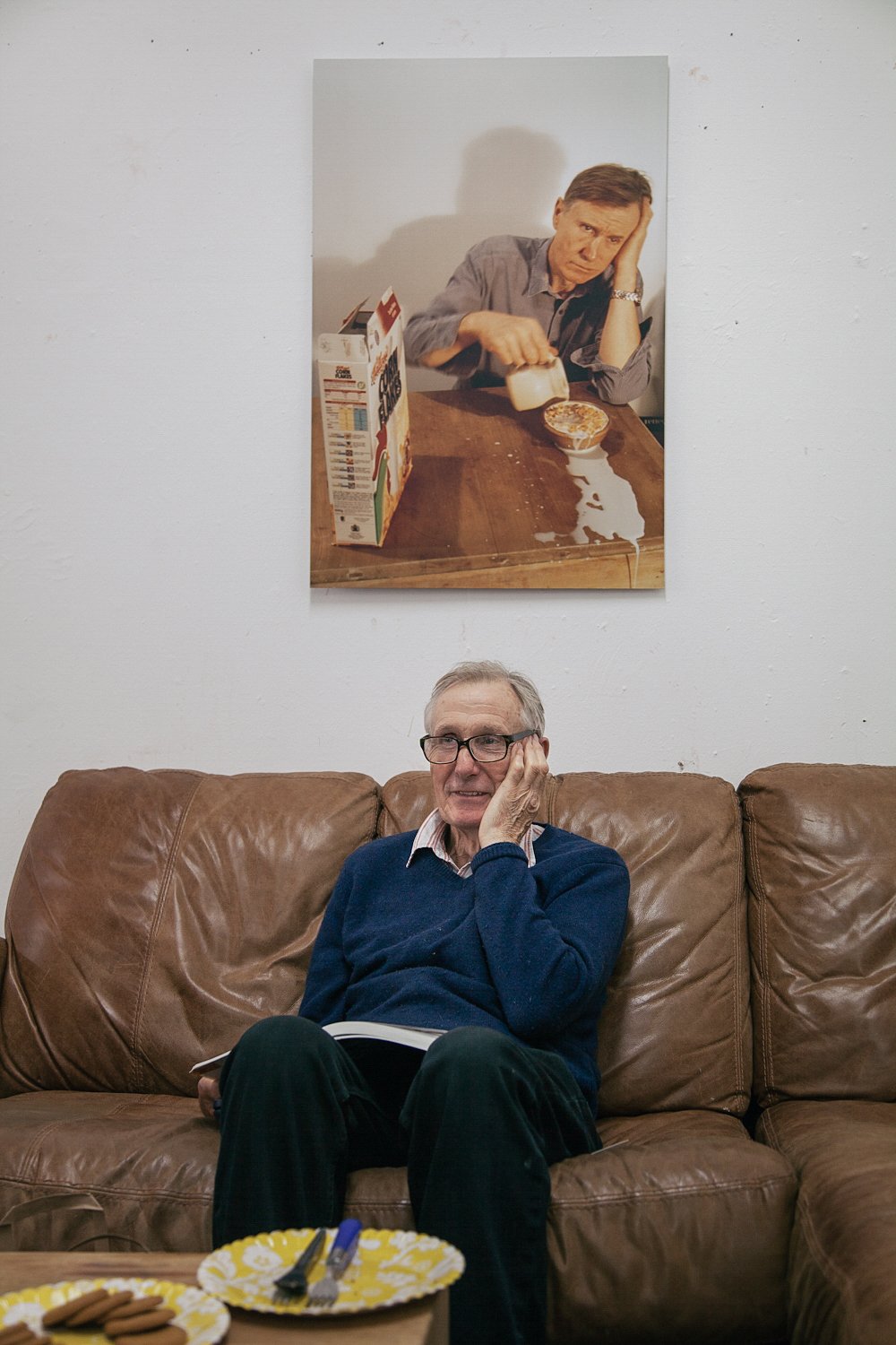 Artist seated on a sofa in a studio at Chisenhale Studios, London, with a framed photograph of him on the wall.