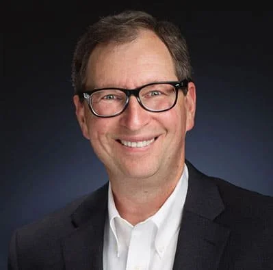A smiling man with glasses wearing a suit jacket and white shirt against a dark background.