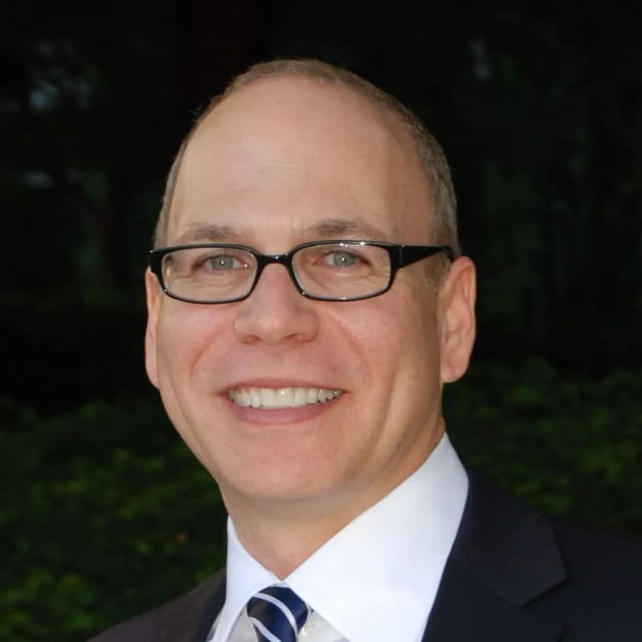 A man with short hair, glasses, and a big smile, wearing a suit, white shirt, and striped tie, outdoors with a dark, blurry background.